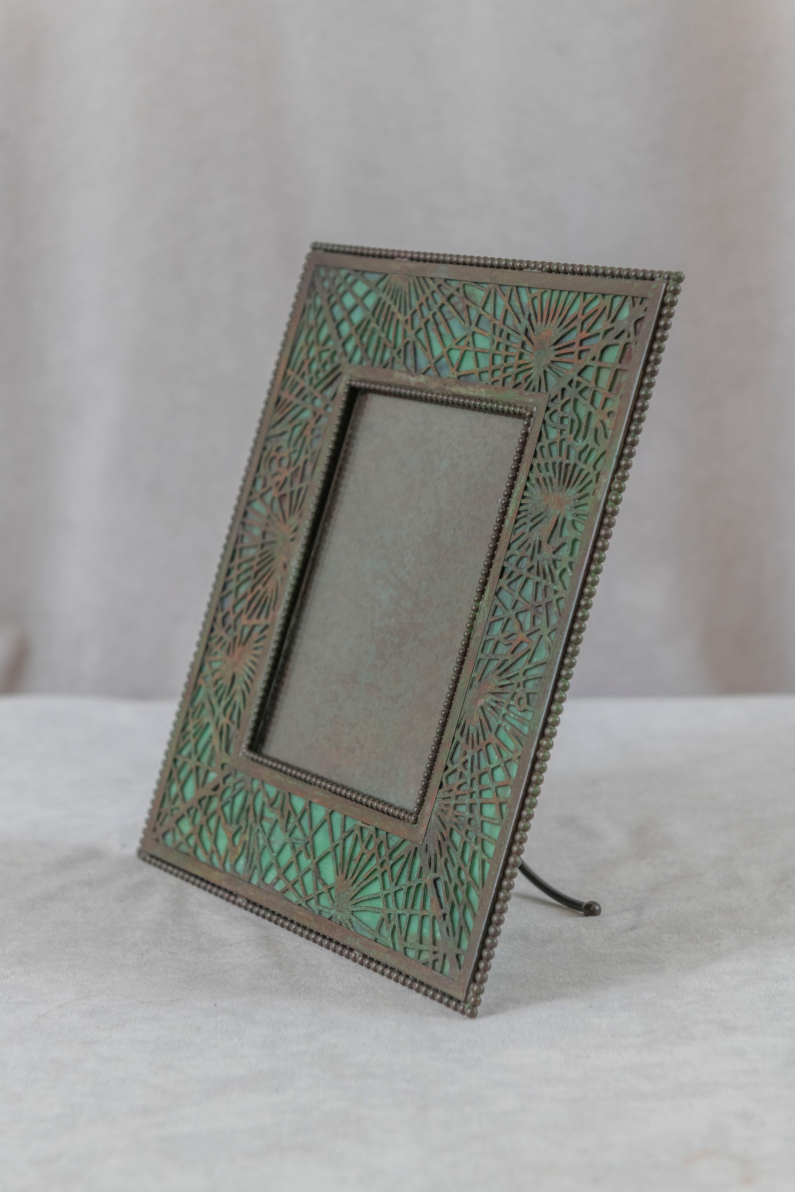  This picture frame was made by the best, Tiffany Studios. Beautiful pine needle design in green patina and colorful glass behind the work. Better glass than normally found on these frames. Properly signed on the pack and all glass intact.
  A real