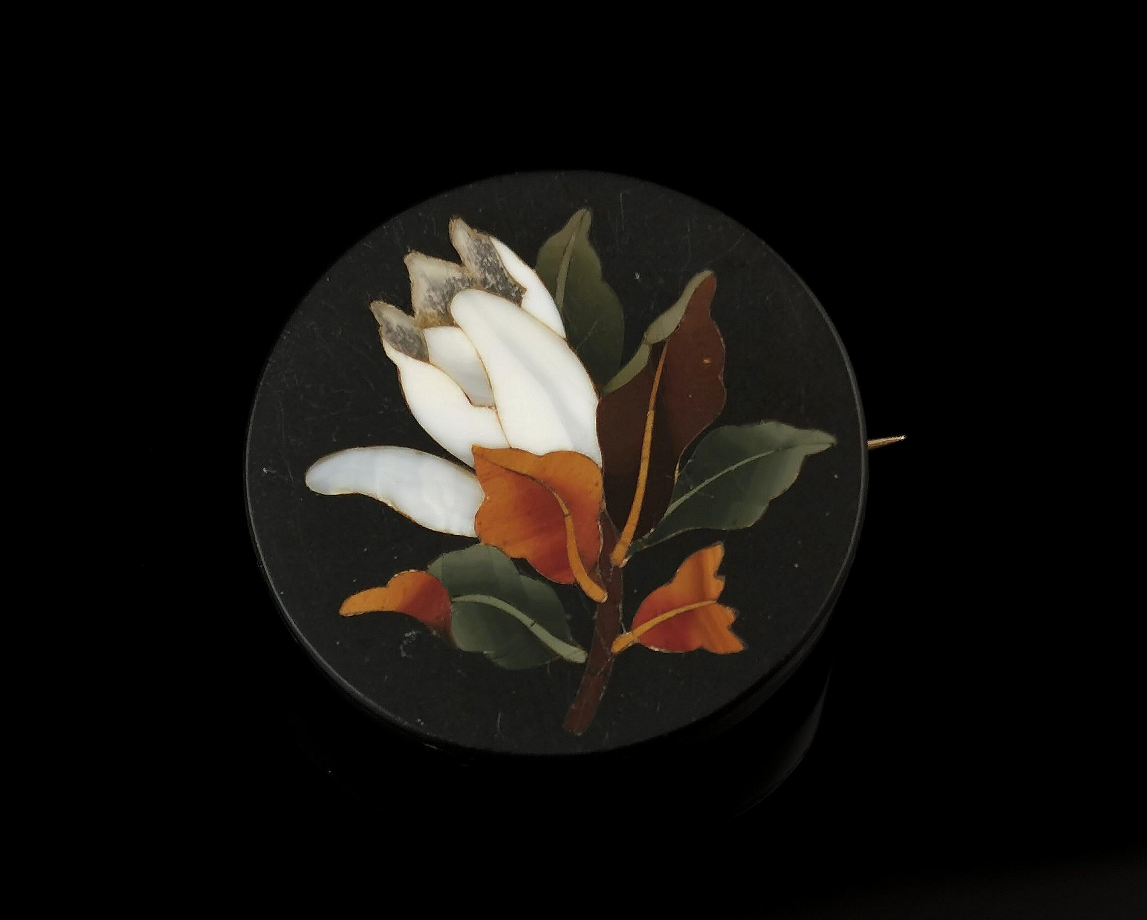 A beautiful antique Pietra Dura flower brooch.

Circular shaped it has a dark slate black ground with hardstone inlay of browns, orange, white and green.

The image depicts a flower and it has a lovely natural colourway.

The brooch has a gilt brass