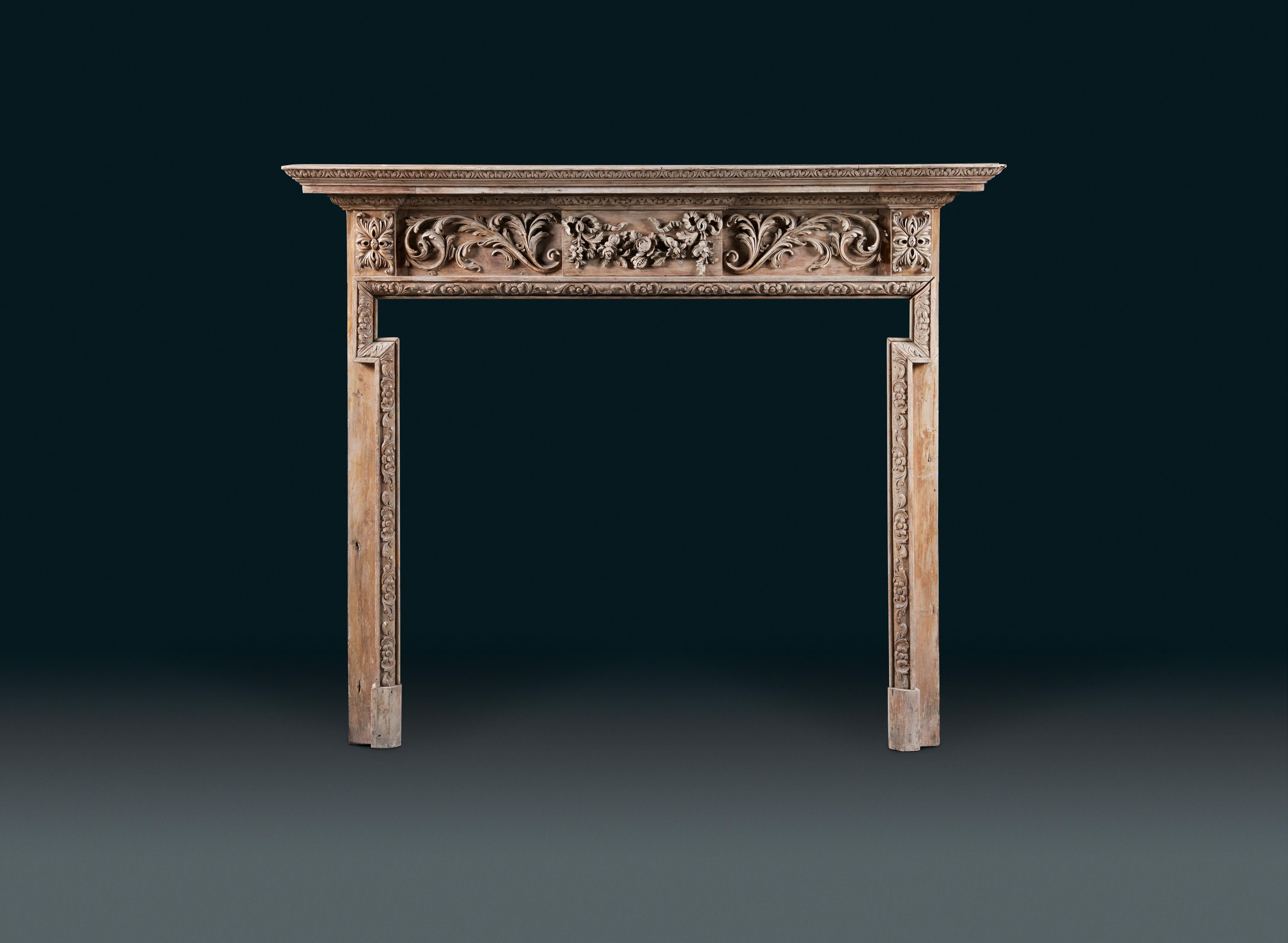 A carved pine fireplace surround of 19th Century date, in an earlier 18th Century style, stripped and waxed; with stepped moulded and decorated shelf, the centre tablet vigorously carved with swags of full open flowers hanging from windblown,