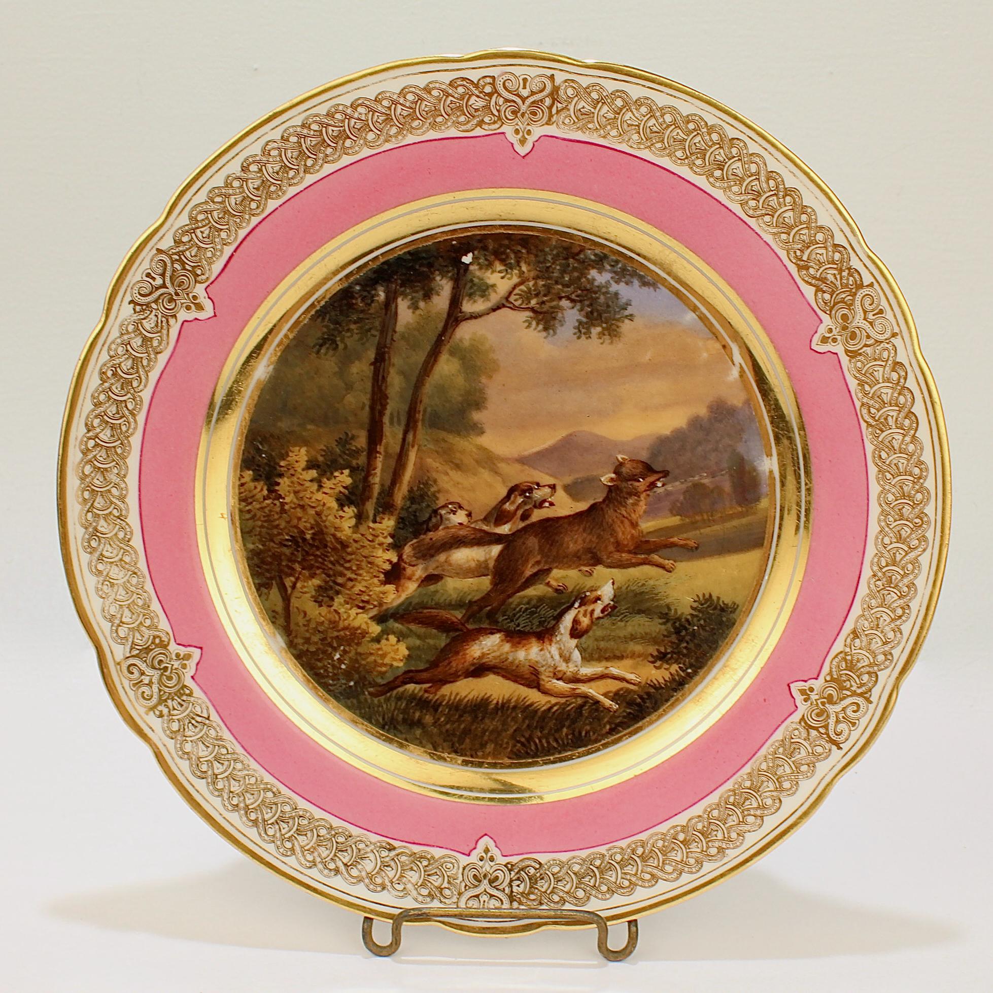 A wonderful antique 19th century Paris porcelain plate.

With a hand painted scene of a fox hunt in a bucolic landscape including a fleeing fox and 3 hounds in chase. 

It has a rich pink border and an elaborate gilt design to the rim and a