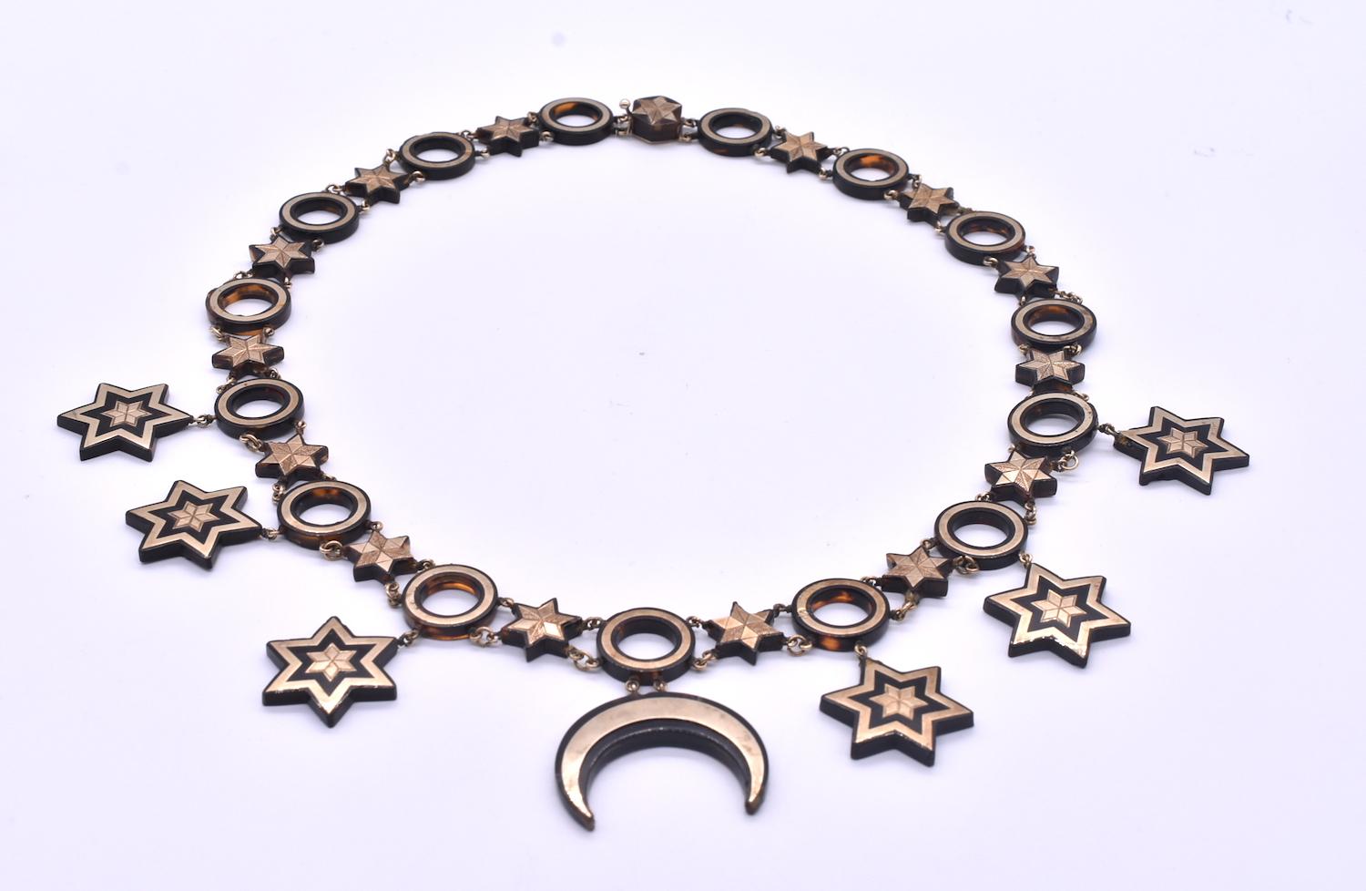 This is an unusual pique necklace with pique star and circle links throughout. From each circle link dangles a larger star pendant, while a single crescent moon pendant takes center stage. The pendants and links are adorned with inlaid gold. 

Pique