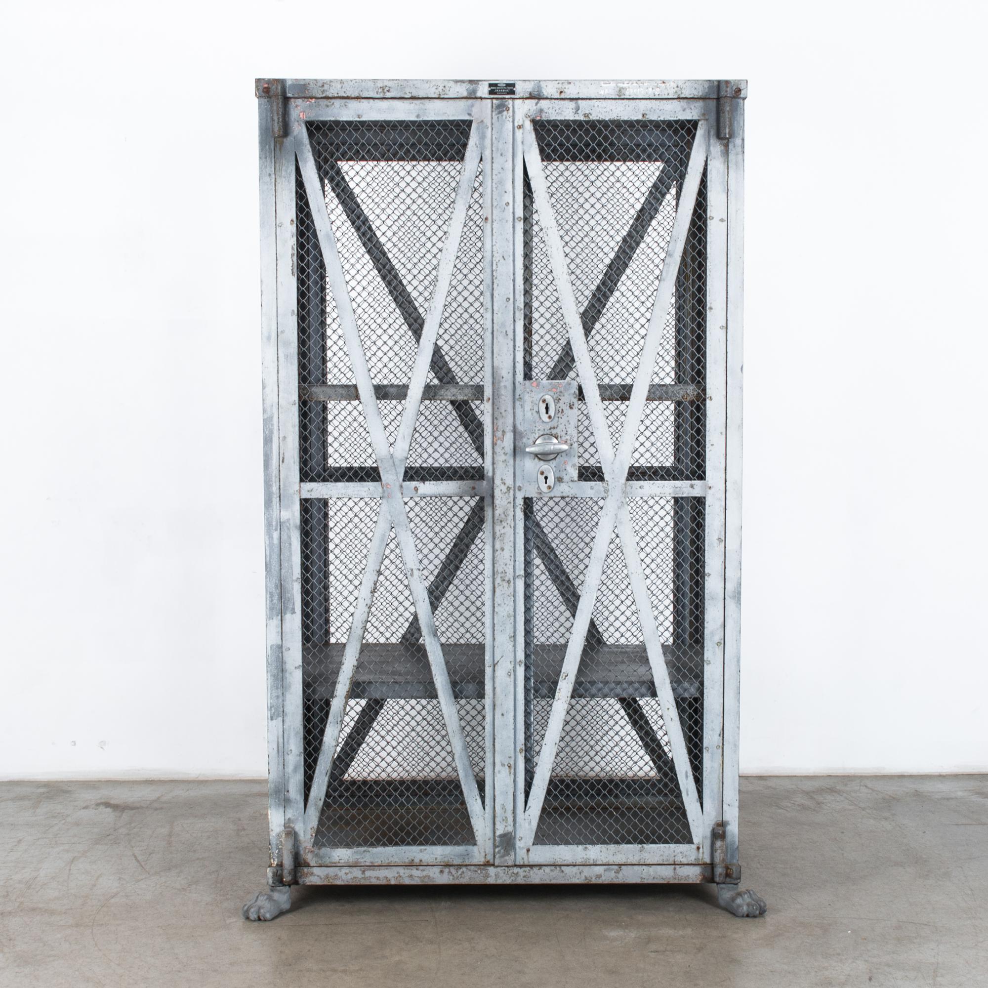This metal safe was produced by “Skarbiec,” circa 1930. A tall cage of wire mesh, reinforced with metal bars, contains two shelves. Claw feet at the base lend a noble touch to the stark silhouette. The striking X-shaped bars and double lock on the