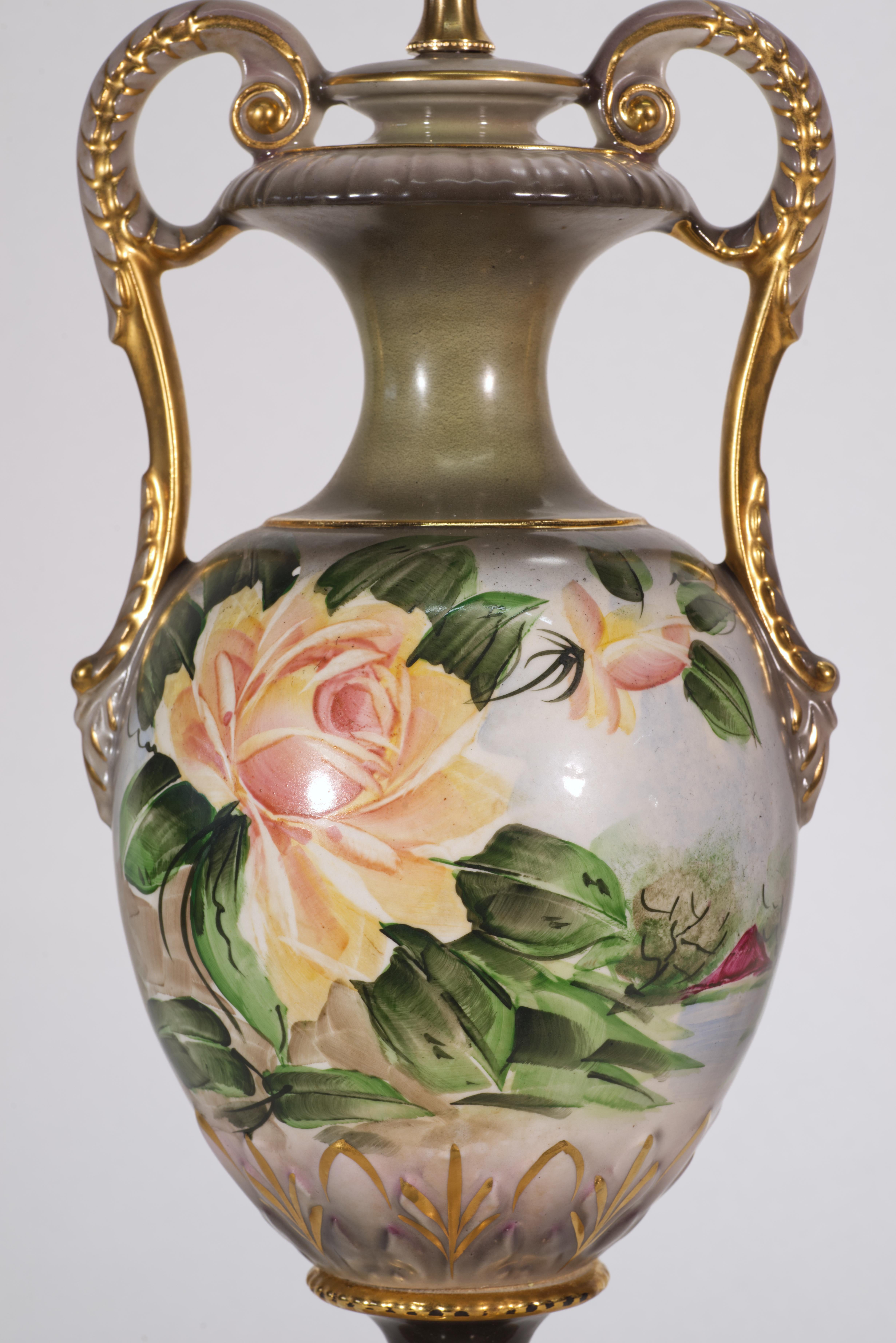La grande lampada antica è caratterizzata da un corpo in porcellana a forma di urna con due maniglie decorative gemelle, dipinte a mano in stile vittoriano con grandi rose inglesi e paesaggi di campagna. I tenui colori giallo crema delle rose sono