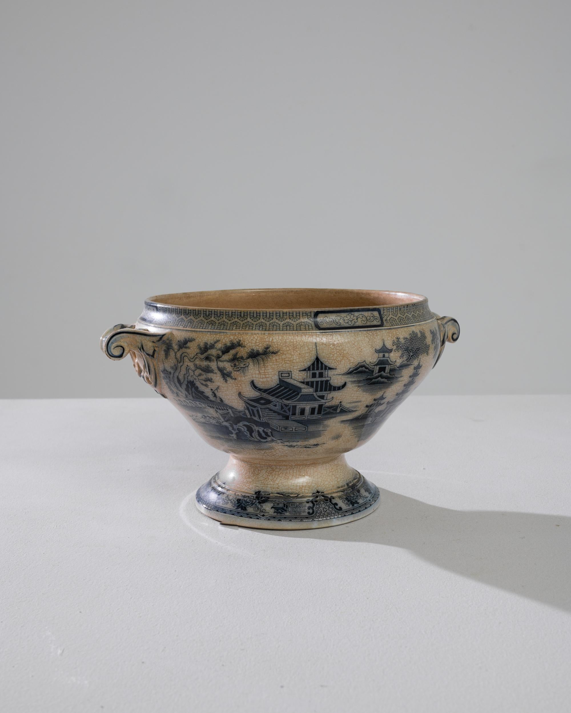 This porcelain tureen was produced in Belgium, circa 1900. The decorated tureen is also adorned with two swirling handles allowing for comfortable use. The geometric blue friezes and charming seaside scene emerge from a textures earth-tone ceramic