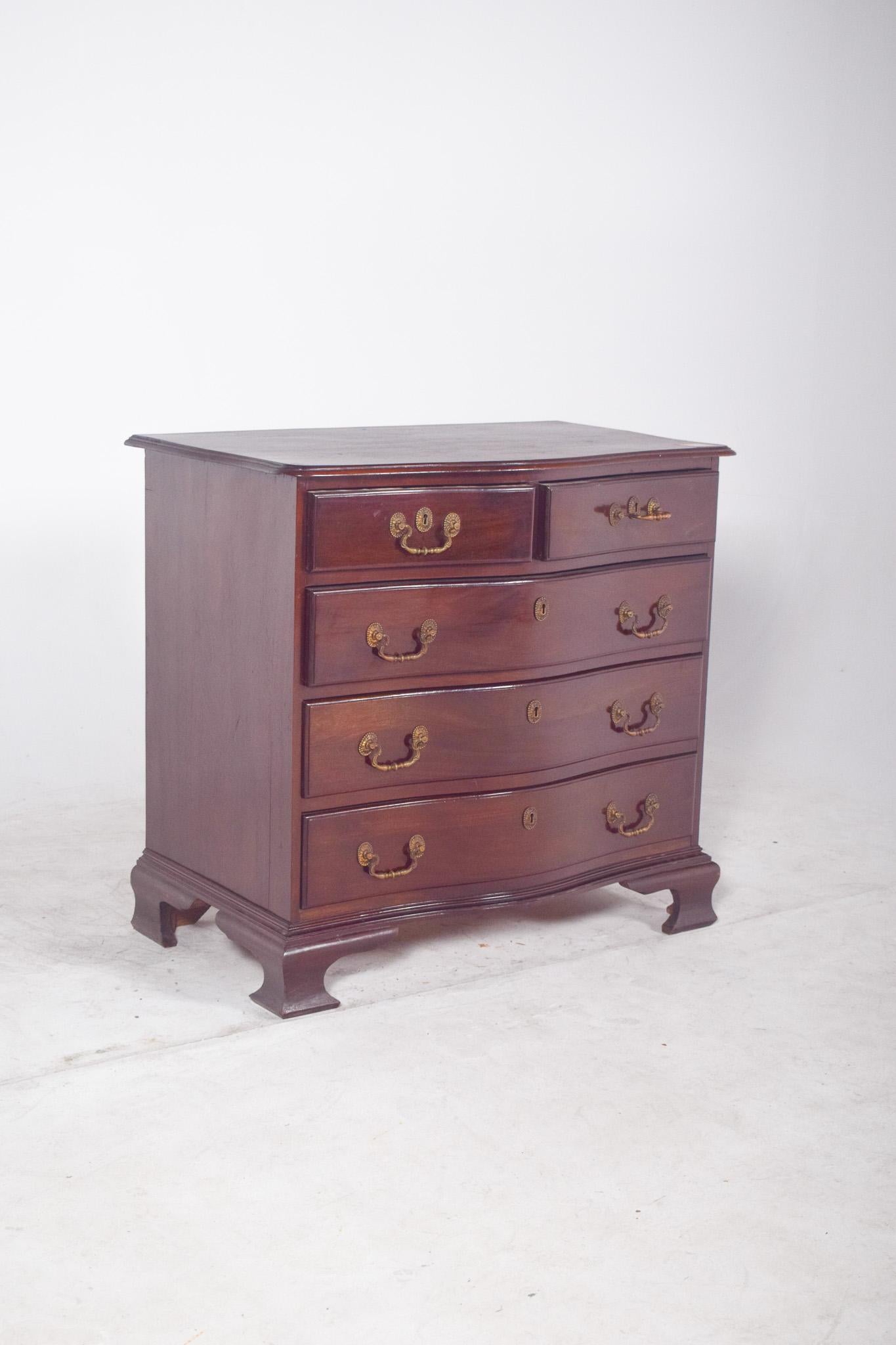 Chippendale-style small chest of drawers. In dark wood, it has five drawers distributed into two small upper drawers and three larger lower drawers, all with hinged metal handles and classically-inspired metal lock mirrors.
The furniture rests on