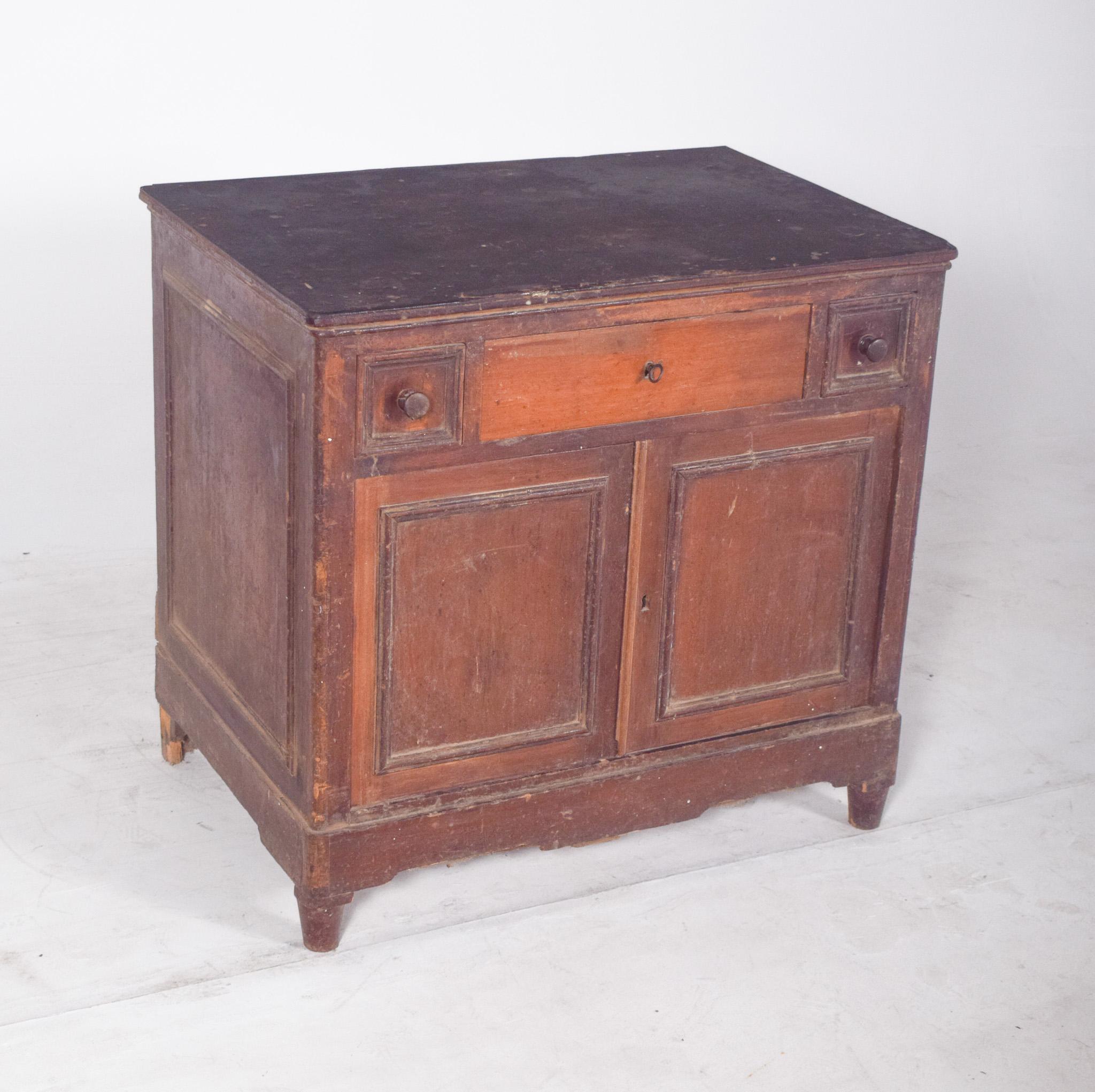 Credenza rustica con due ante e tre cassetti. In legno scuro, con due ante inferiori che si aprono su un ampio spazio interno con un ripiano centrale. Sopra, un cassetto centrale più grande affiancato da due piccoli cassetti quadrati, ciascuno con