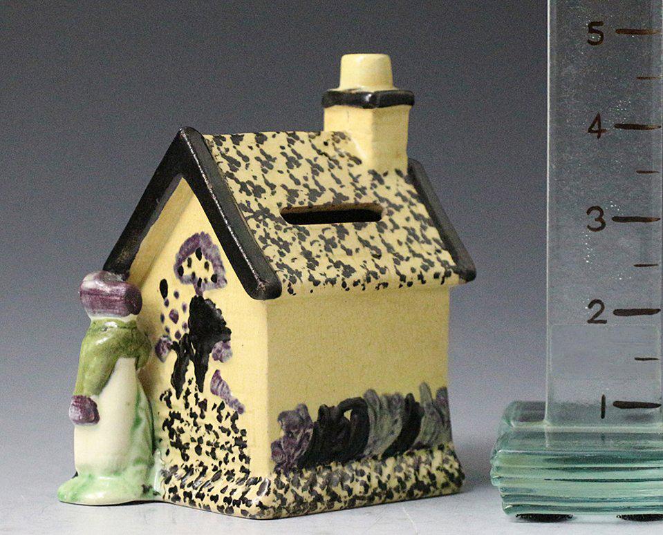 A charmingly naive Prattware colored money box bank in the form of a cottage flanked by male and female figures the head of a child looks out from the upper central window. The sides of the cottage are decorated in relief with a flowering tree there