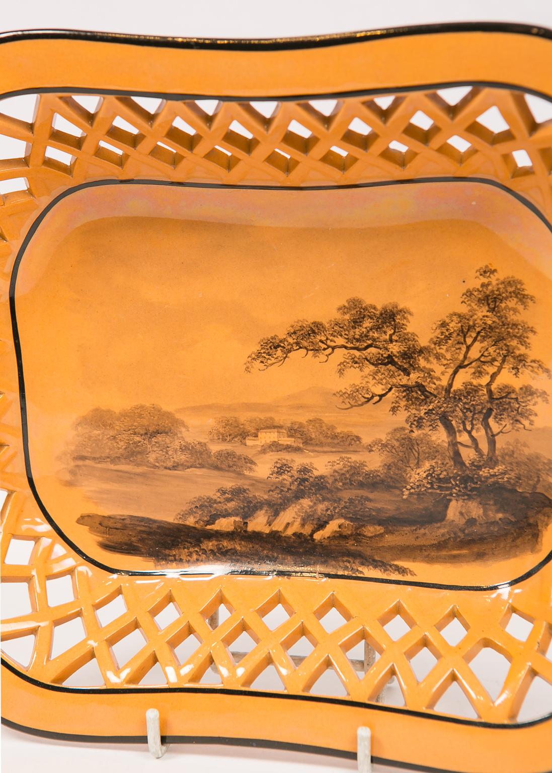 We are pleased to offer this antique pottery pierced basket painted with a country scene on an orange ground.
Printed in grisaille the scene shows an English landscape with a manor house in the background. The piercings are shaped as a web of