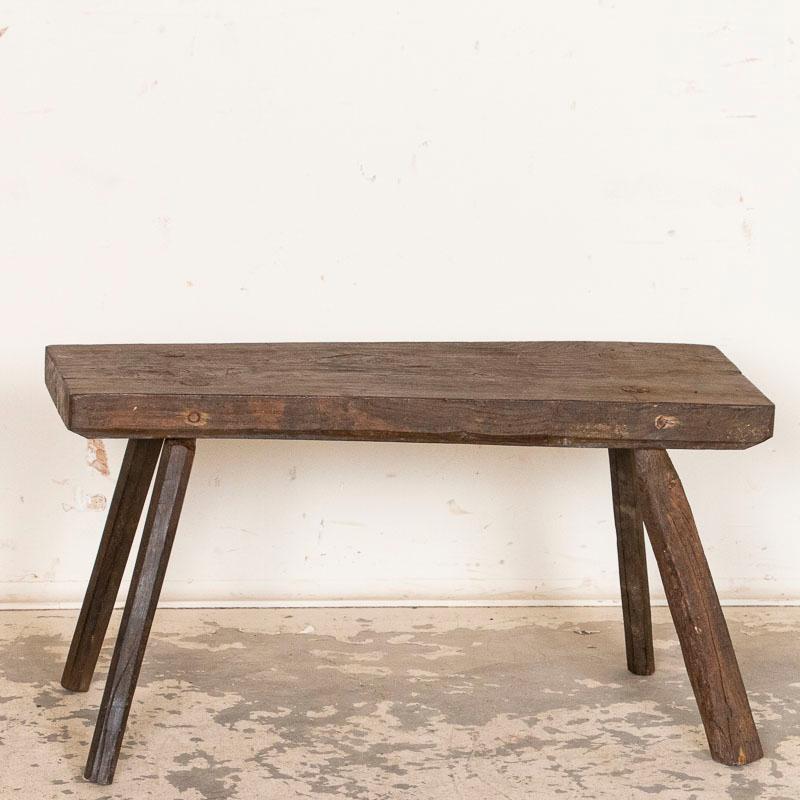 The rustic appeal of this primitive stool or small bench comes from the wood itself, greatly distressed over time leaving a dark, deep patina. The age-related cracks, stains, and scratches all add to its character. The splay legs with pegs showing