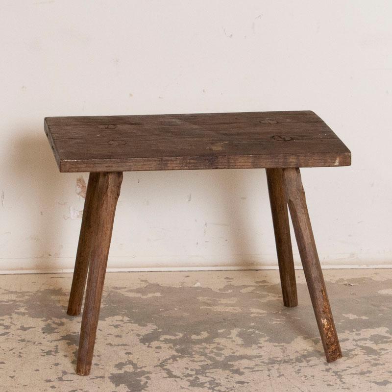 The rustic appeal of this primitive stool or small bench comes from the wood itself, greatly distressed over time leaving a dark, deep patina. The age-related cracks, stains, and scratches all add to its character. The splay legs with pegs showing