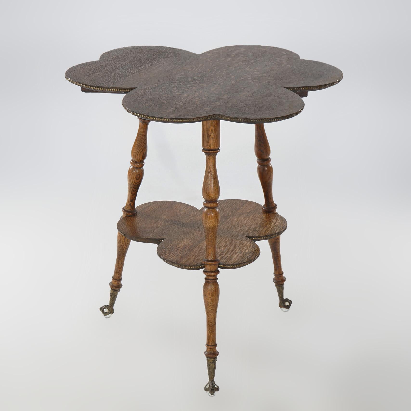 An antique parlor table offers quarter sawn oak construction with top in clover leaf form and having brass bead trimming over matching lower shelf, raised on turned legs terminating in brass claw and glass ball feet, circa 1900

Measures - 29.5