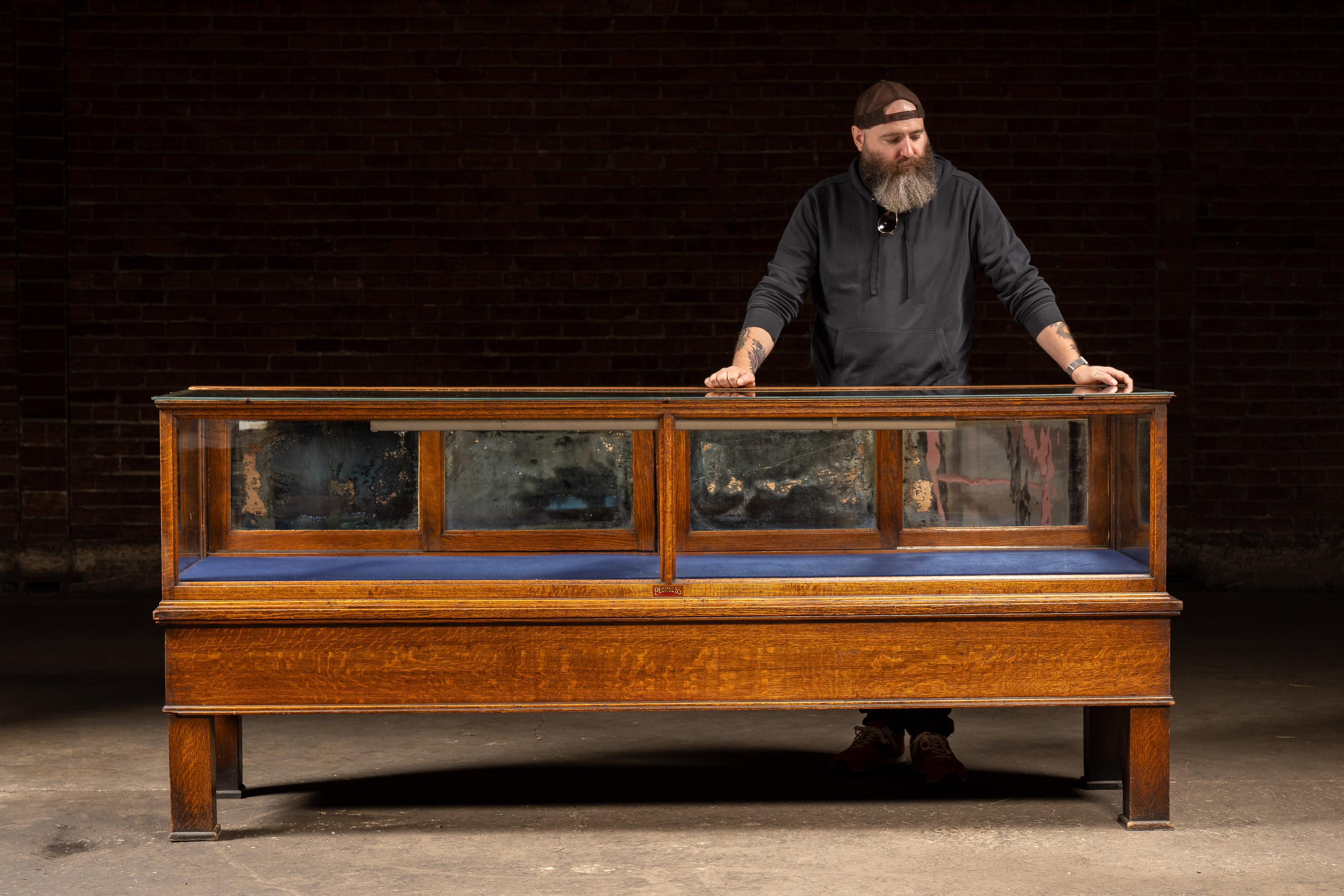 c. 1910er-1920er Jahre; Jac. Lederer, Inc. - Chicago, IL

Große, viertelgesägte Eichenholzvitrine von Peerless Store Fixtures mit gerahmter Glasplatte und -front, verspiegelten Innenpaneelen und reichem Goldfinish. Die Vitrine behält ihr originales