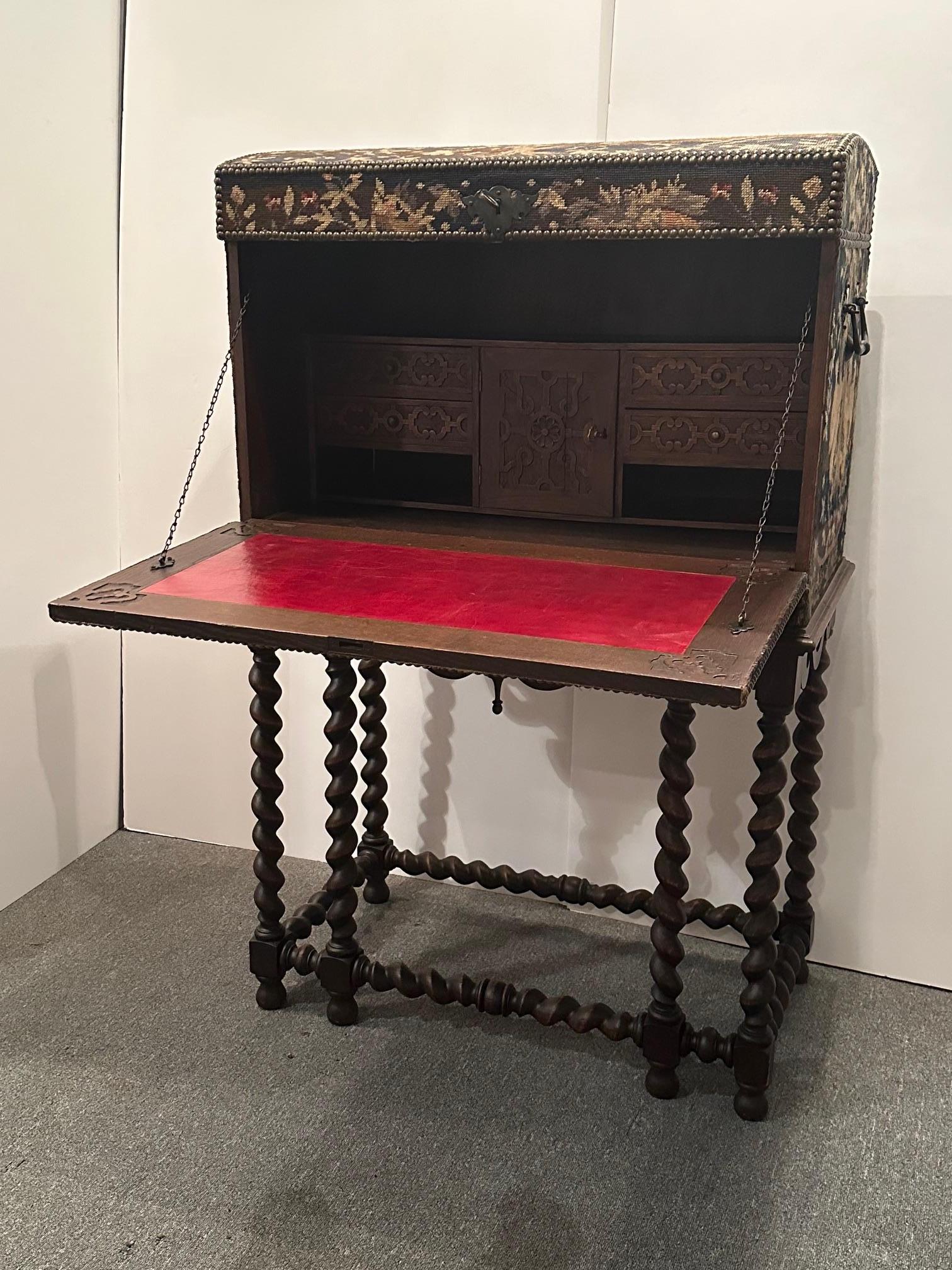 Unusual and magnificent trunk shaped desk covered in gorgeous rich tapestry needlepoint and brass nailheads.  The top piece opens to reveal a drop down writing surface with red leather.  The dark stained oak base has handsome barely twist legs. 
