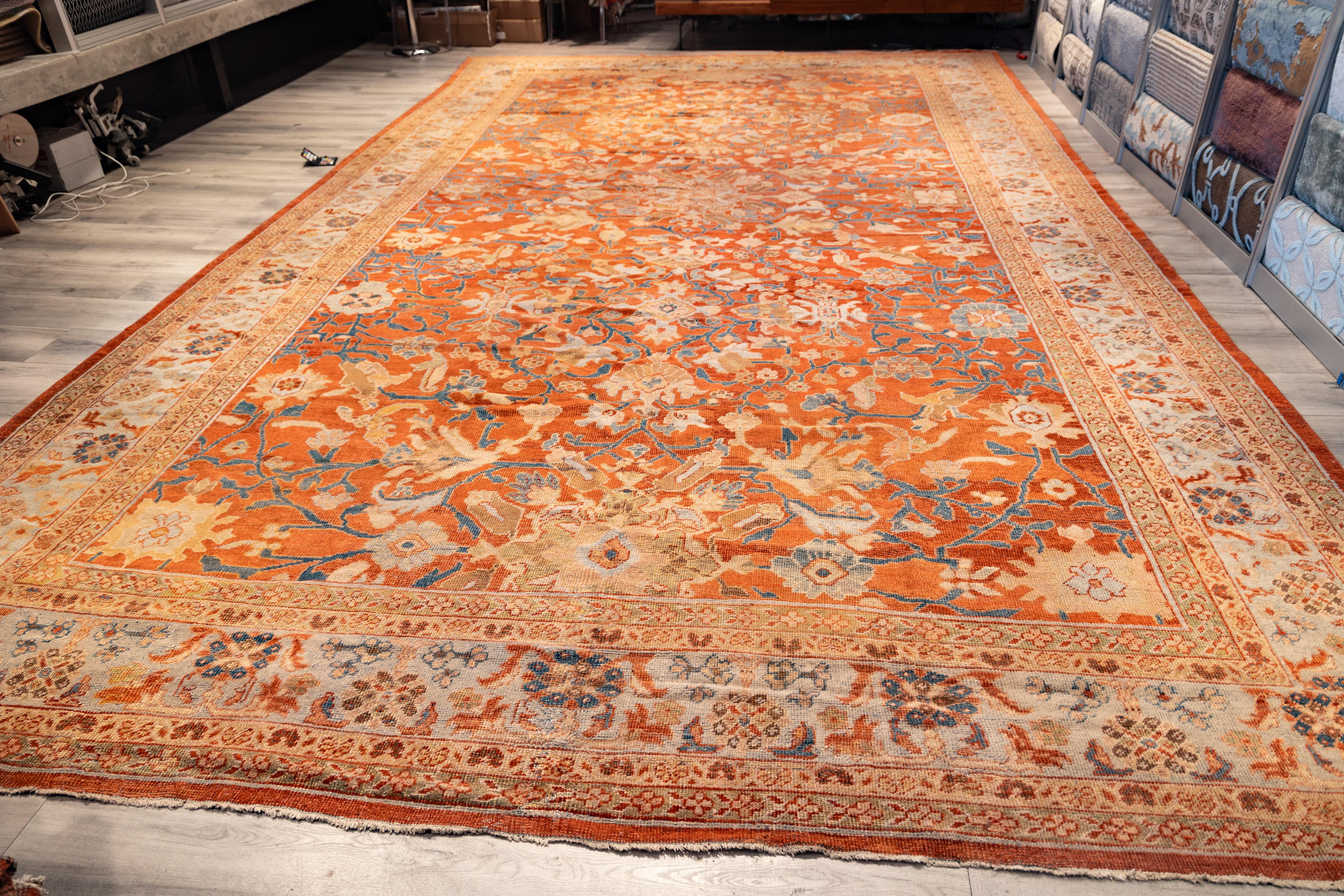 Large Sultanabad rug features a soft red field filled with floral vines, palmettes, and rosettes in shades of ivory, light blue, gold, and coral. The wide ivory border frames the design with a repeating pattern of flowers and curling leaves. The