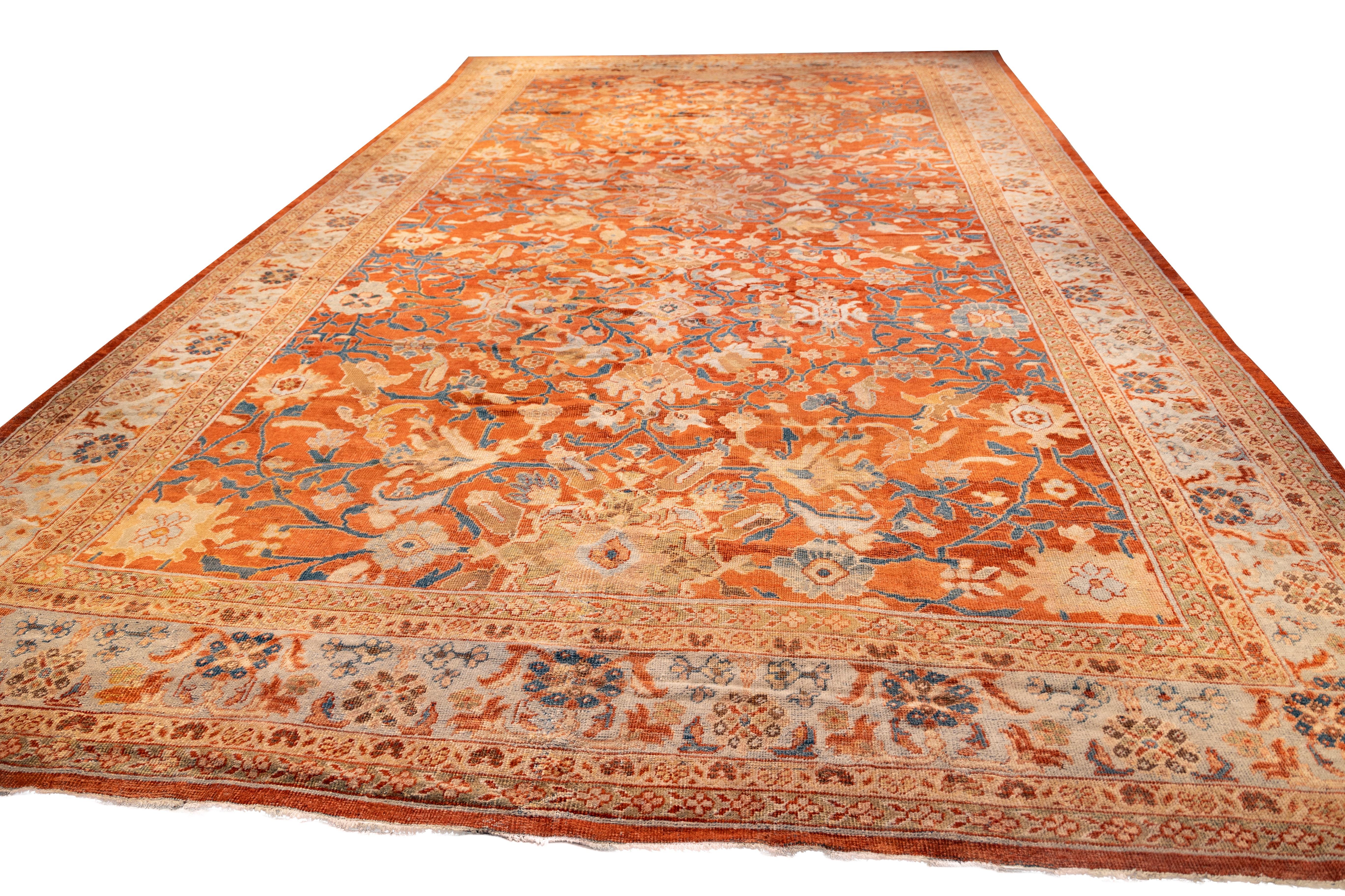 Large Sultanabad rug features a soft red field filled with floral vines, palmettes, and rosettes in shades of ivory, light blue, gold, and coral. The wide ivory border frames the design with a repeating pattern of flowers and curling leaves. The