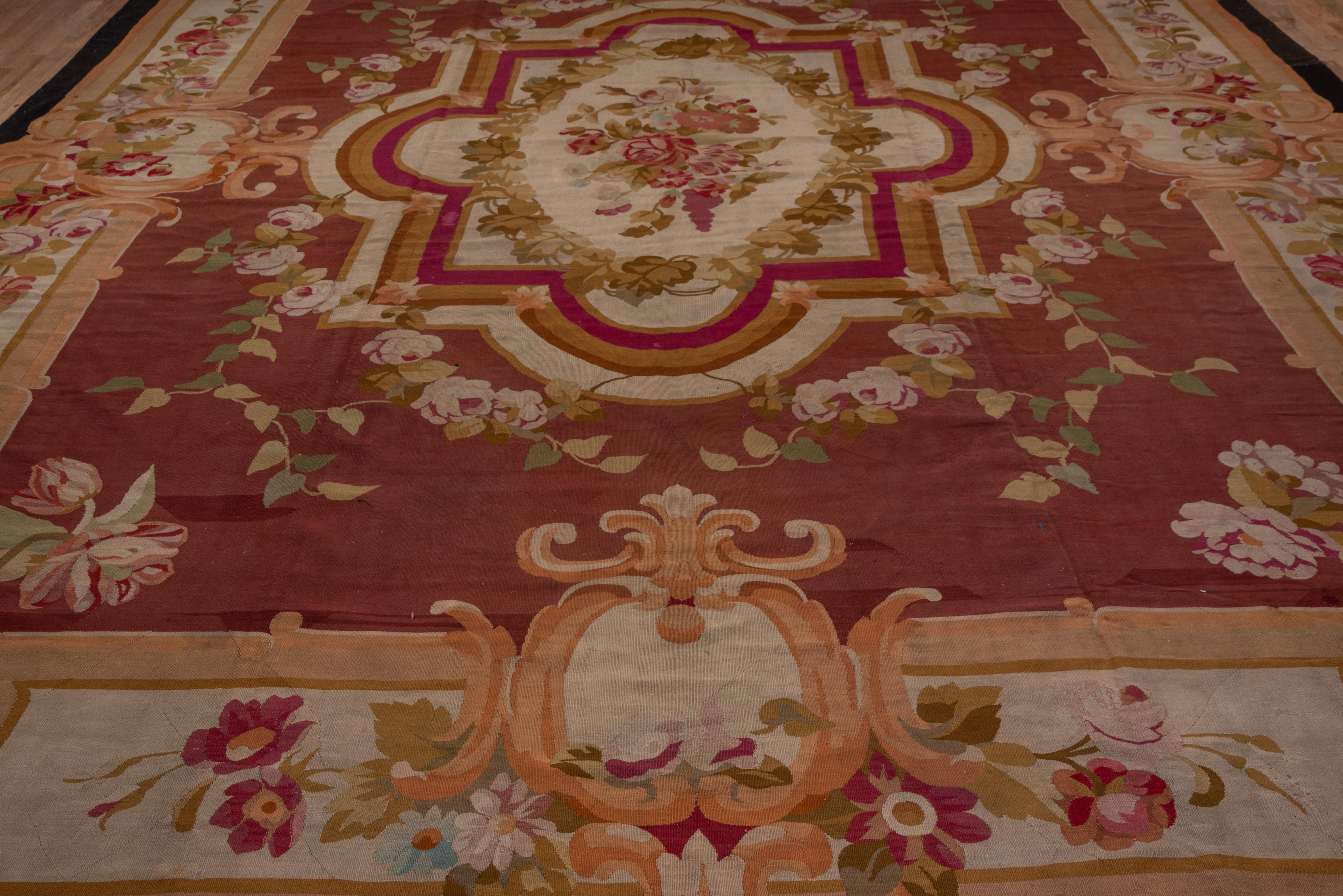 The rich red field displays a stepped and rounded octogramme medallion enclosing an ivory oval floral wreath and a central rose bouquet. Floral sprays are laid out symmetrically all around. The ivory border of this antique French tapestry-woven