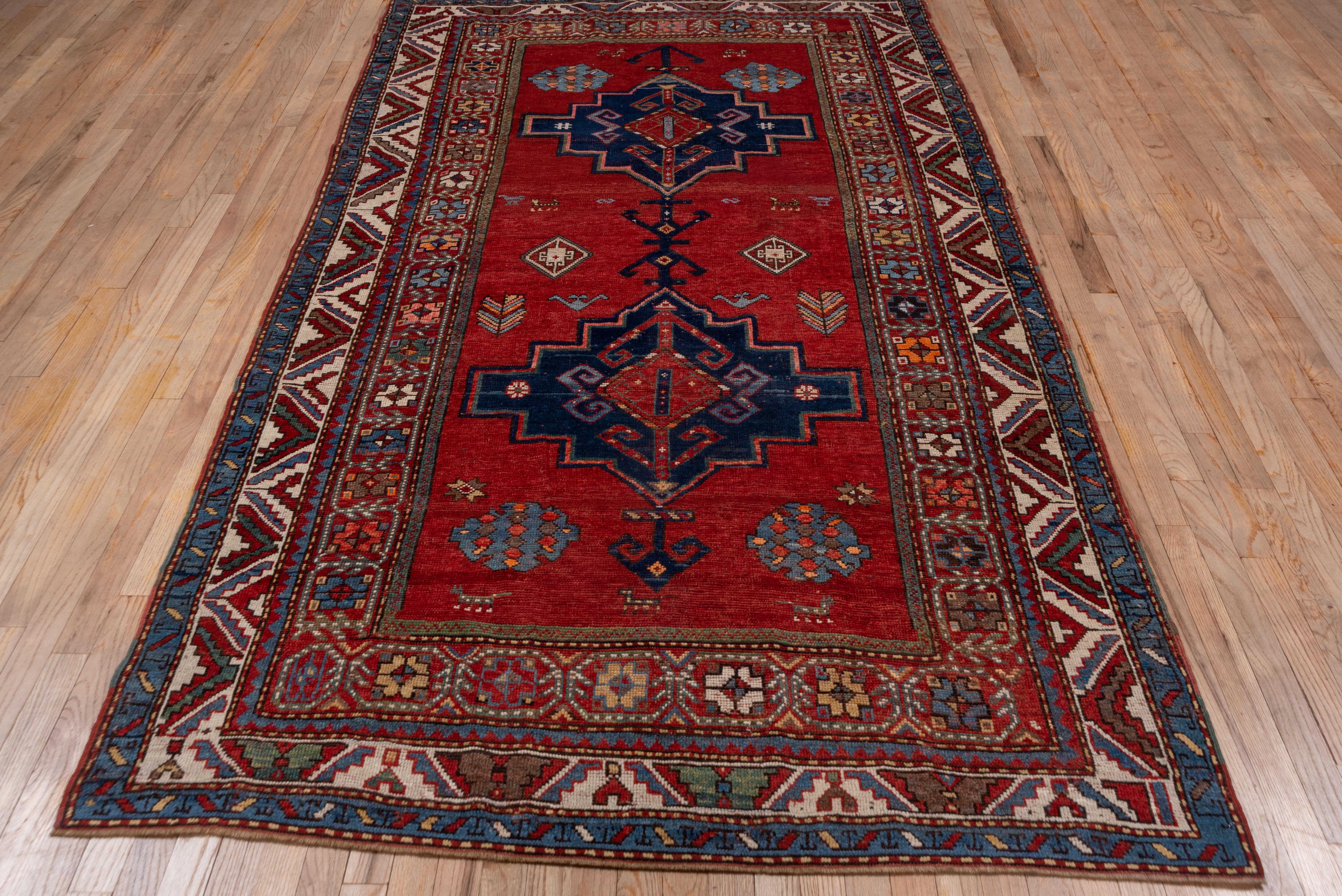 The saturated madder red field of this rug displays two large stepped and pointed royal blue medallions with red voluted centres. Scattered around are tiny animals, complex rosettes, stars, feathery plants and dark brown 