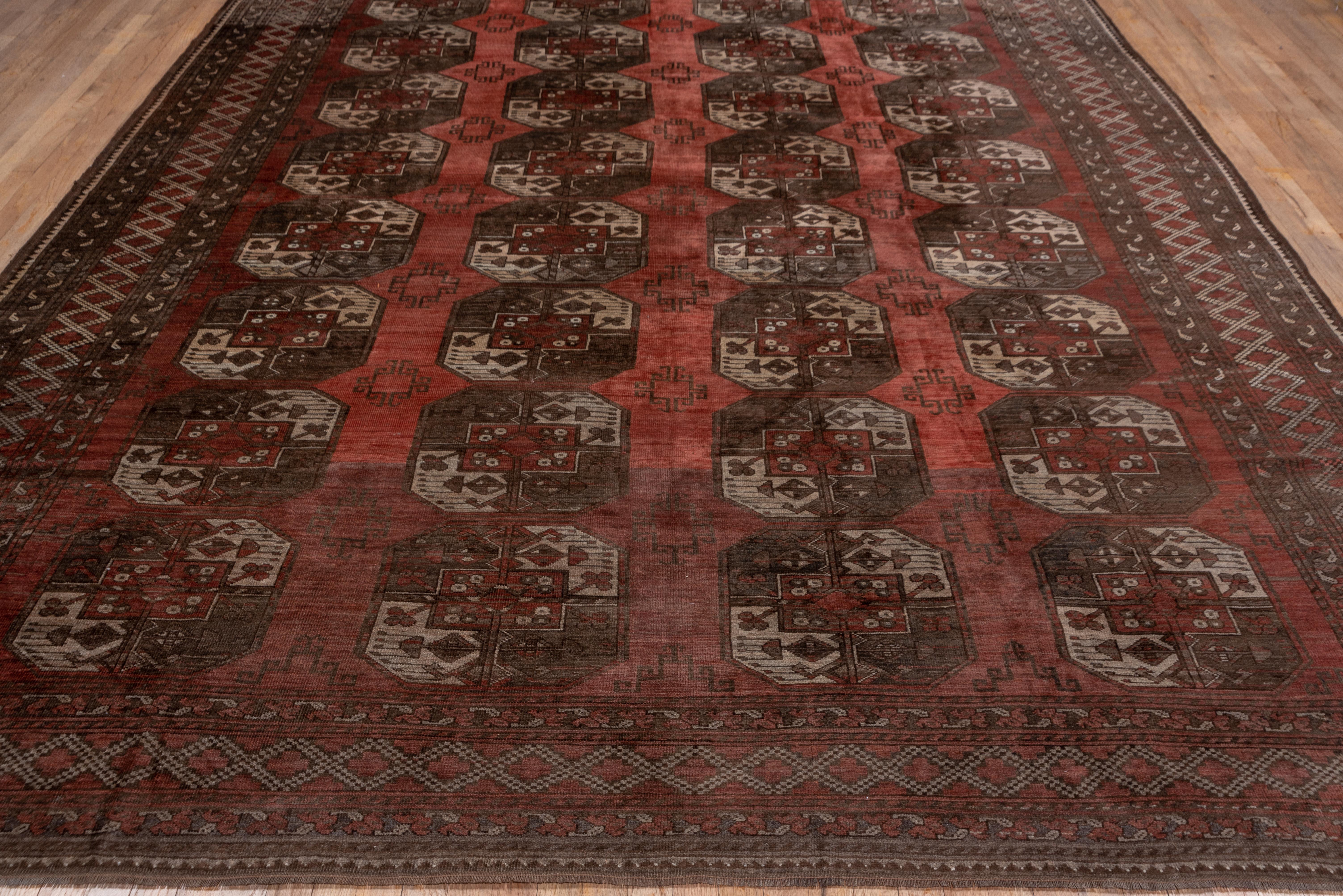 The lightly abrashed madder brick field on this carpet displays four columns of eight characteristic quartered octagonal tribal guls with green andburnt buff sections. The minor intermediate guls fade into the background. The green ground minors