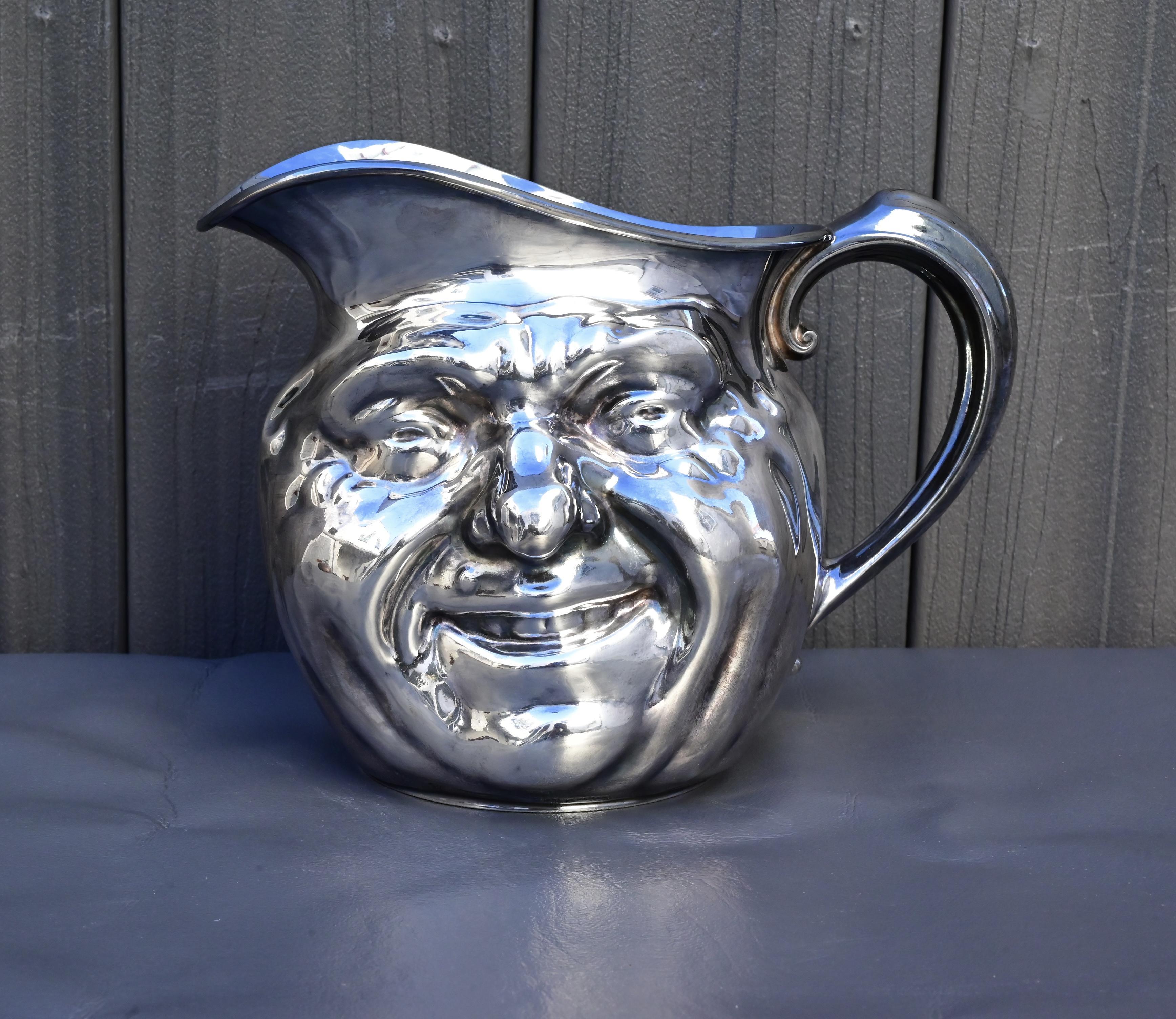 This charming antique silver-plated pitcher by Reed & Barton features a wonderfully expressive face motif design, often referred to as a “Jolly Face” or “Sunny Jim.” Measuring approximately 9 inches across from the handle to the spout and 6¾ inches
