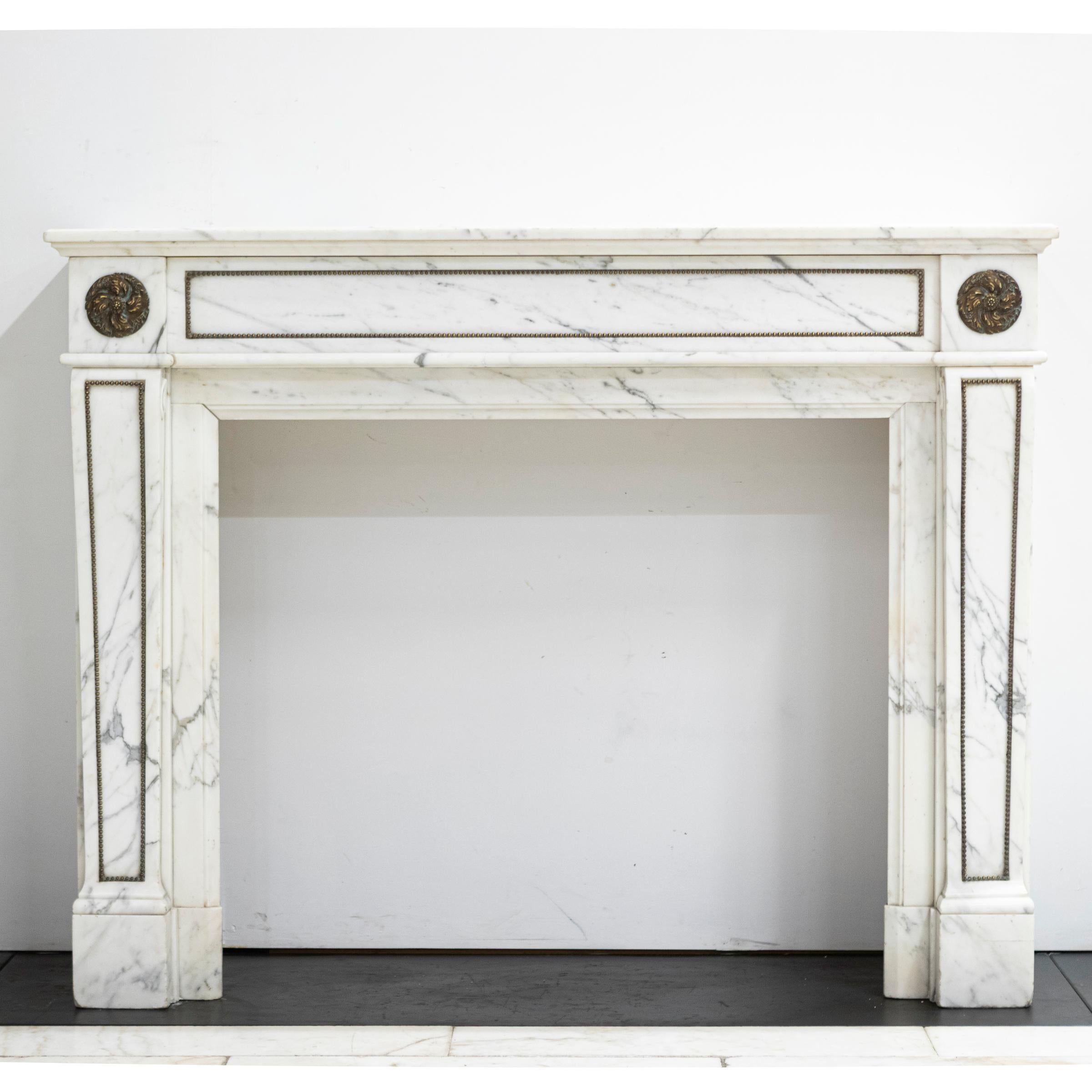 This antique Louis XVI fireplace surround has been finely carved from Italian Arabasque marble that provides high contrast veining. Flower brass ormolu surmount the jambs. Brass mounts and studs line the panels of the frieze and