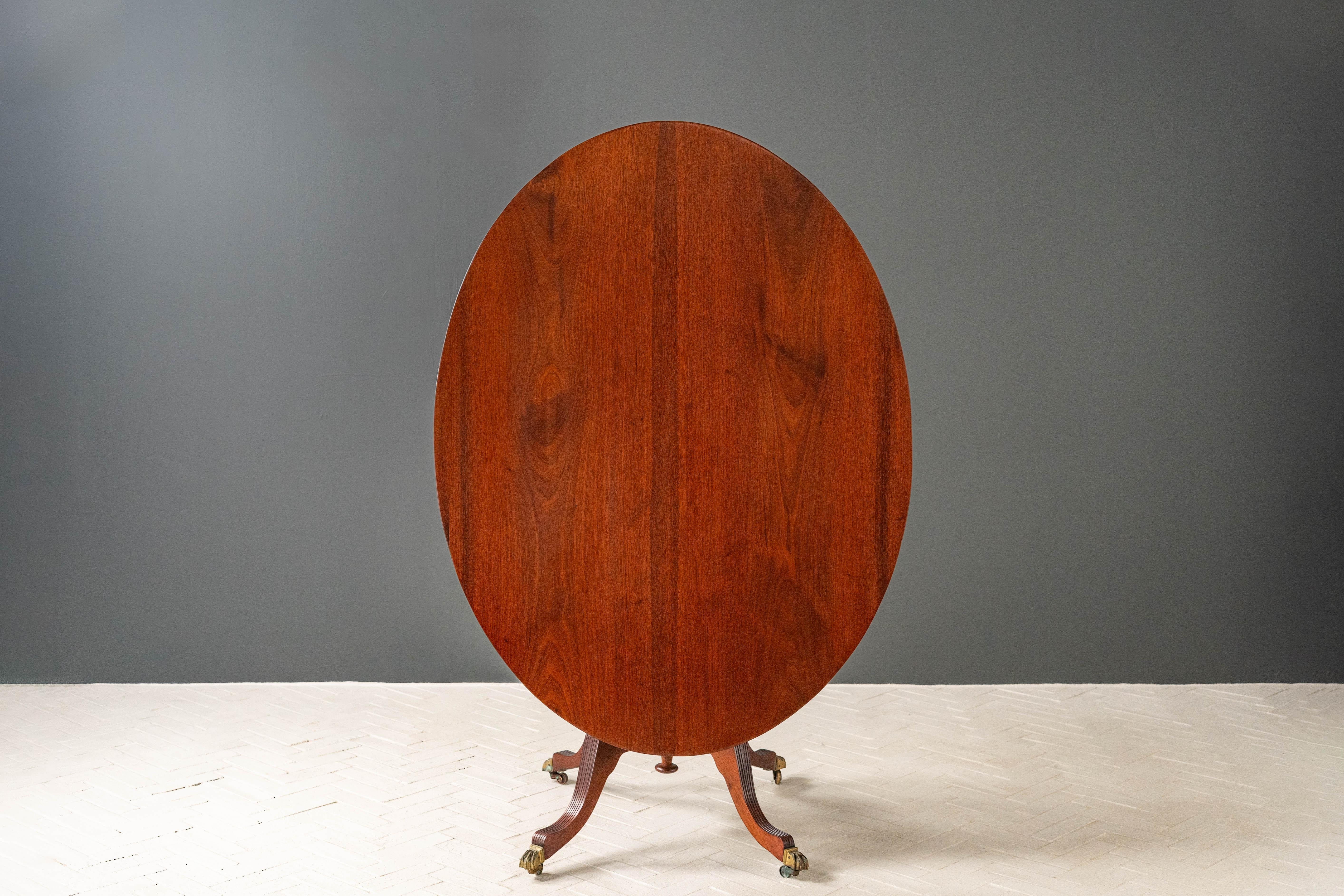 Jamaica, ca. early 19th century. An elegant Regency tilt-top breakfast table. Features an oval top constructed of solid bookmatched mahogany with a turned pedestal base. Four curved legs with carved detailing are supported by brass lion paw feet