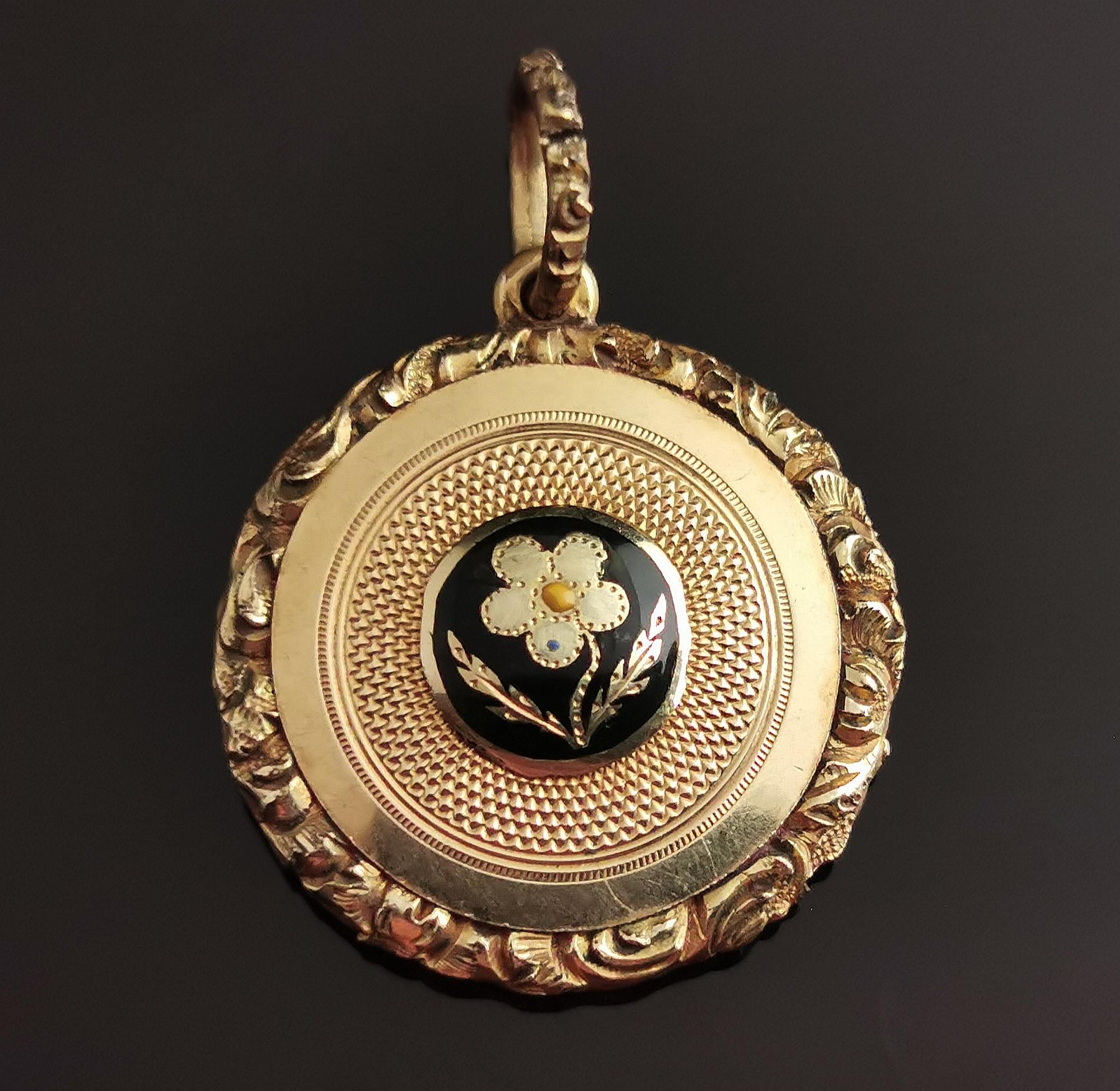 A beautiful antique Regency era mourning locket pendant.

It has an elaborate chased and engraved frame and bale and a gold panel front with black enamel flower, the reverse has a glazed compartment containing a lock of light brown hair secured with