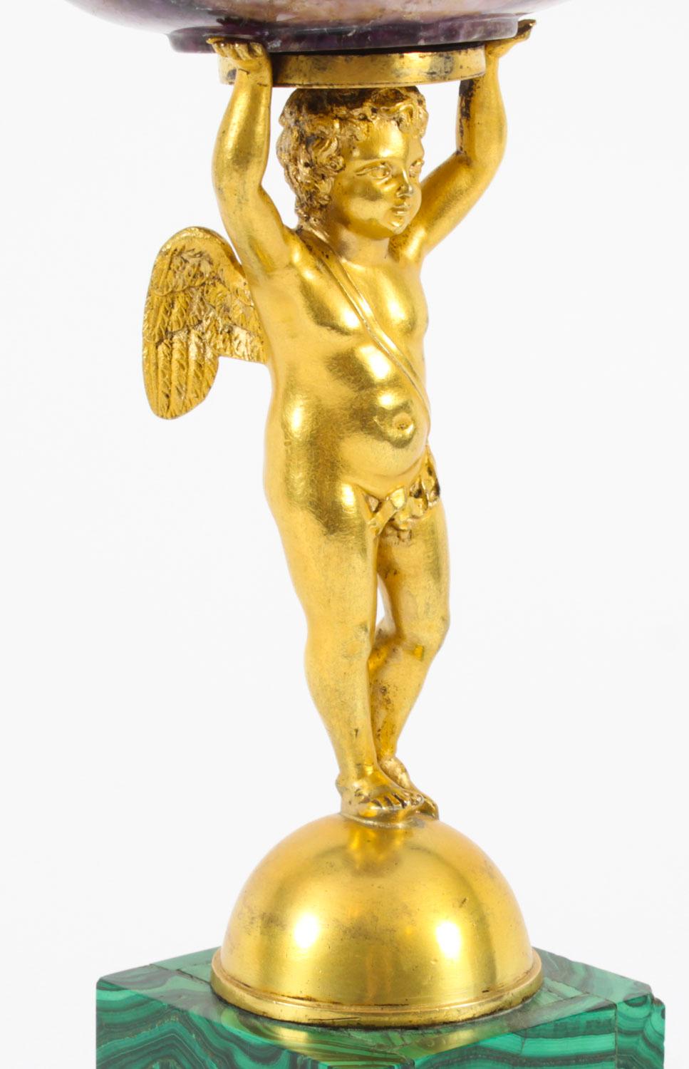 Si tratta di un'impressionante e altamente decorativa tazza del periodo Regency in ormolu, malachite e Blue John,  Datazione circa 1820.

Presenta  un cherubino alato in ormù che regge una tazza Blue John su una base a gradini in