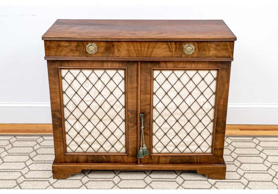 An elegant grid Front cabinet with handsome graining, fine traditional form and elaborate hexagonal brass drawer pulls. Two doors open to shelving. Doors with fabric panels with metallic lattice work. It has a key to lock. Above is a long drawer