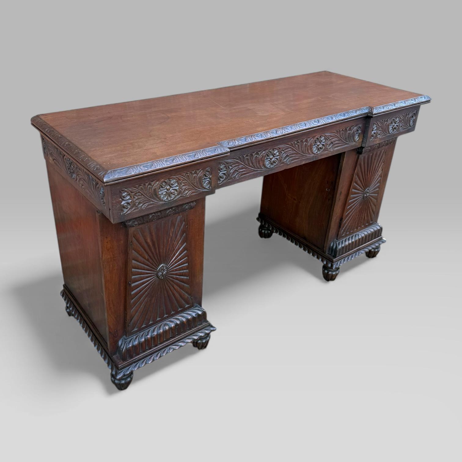 A early 19th century William IV / late Regency period Anglo-Indian inverted breakfront pedestal sideboard crafted in solid padouk with an excellent patina and colour. Likely made in Bombay or Southern India, the piece reflects the skilled carving