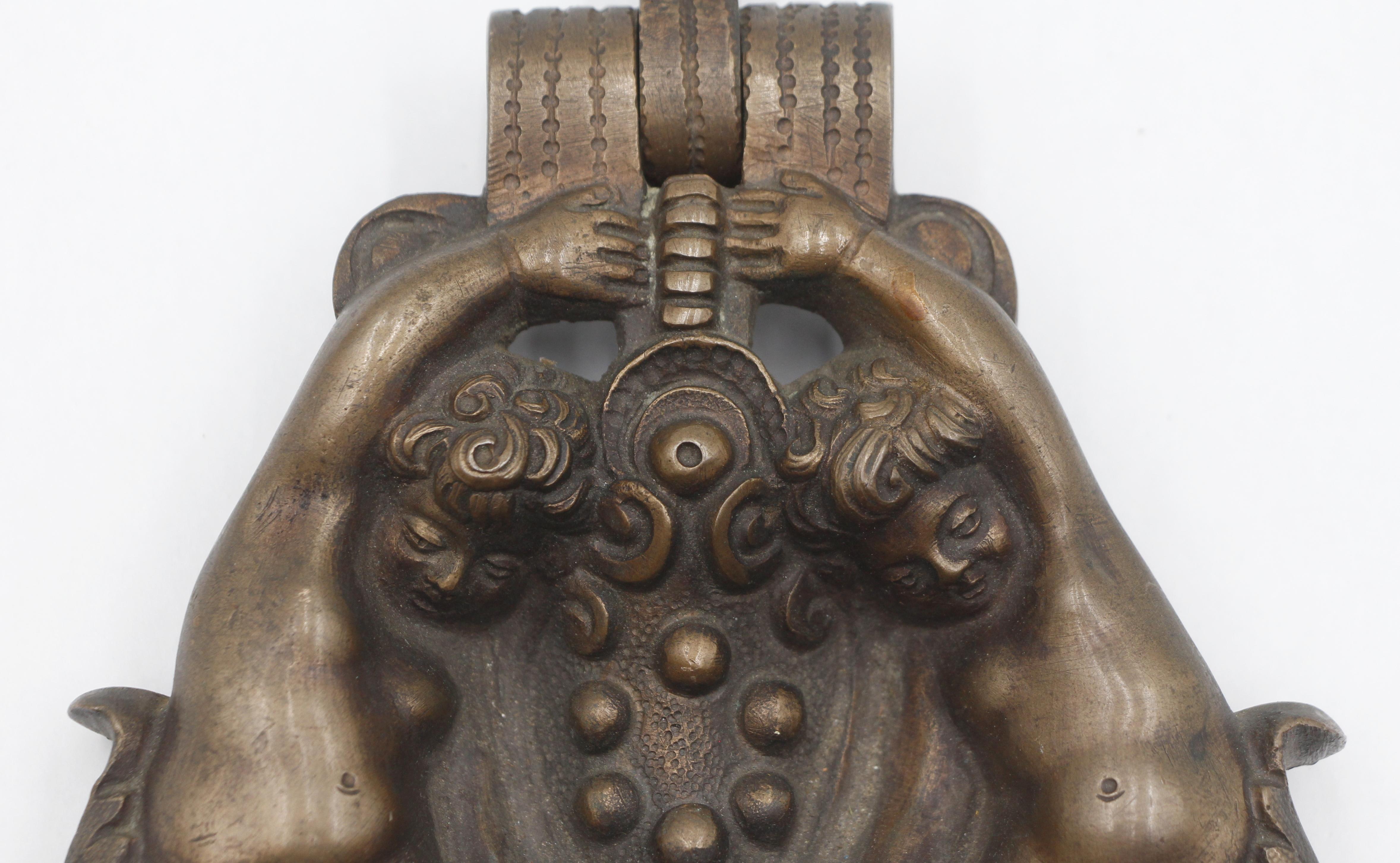 Renaissance style door knocker featuring two cherubic figures holding a central element, possibly a shield or a decorative motif. It is made of cast bronze with a dark patina. The original mounting nut is not included. Good condition with