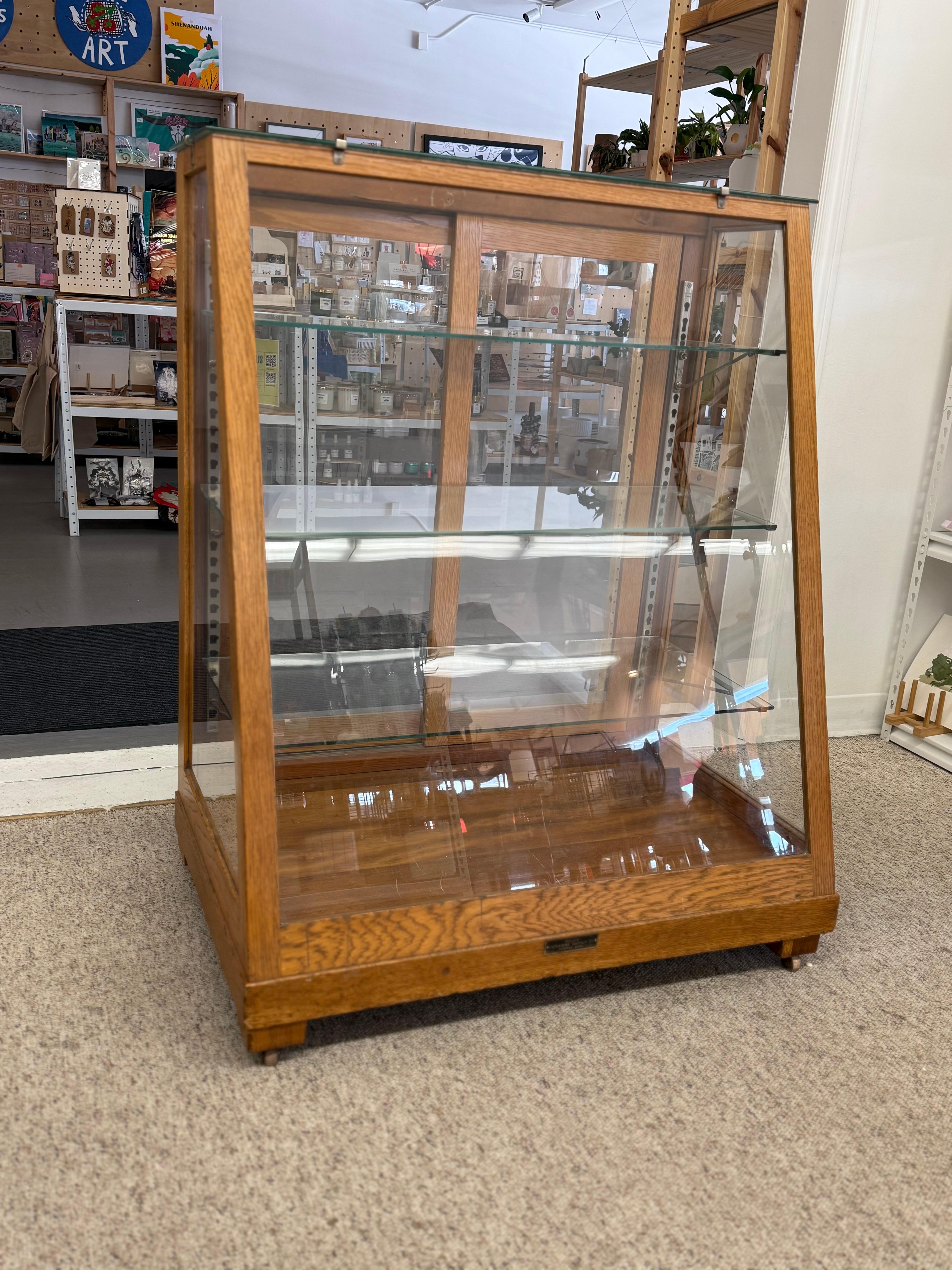 The cabinet is a vintage display case made of oak wood with a rich, natural finish. It features a slanted, A-frame design with a glass top and three glass shelves inside, all enclosed by glass panels on the sides and front with sliding doors. The
