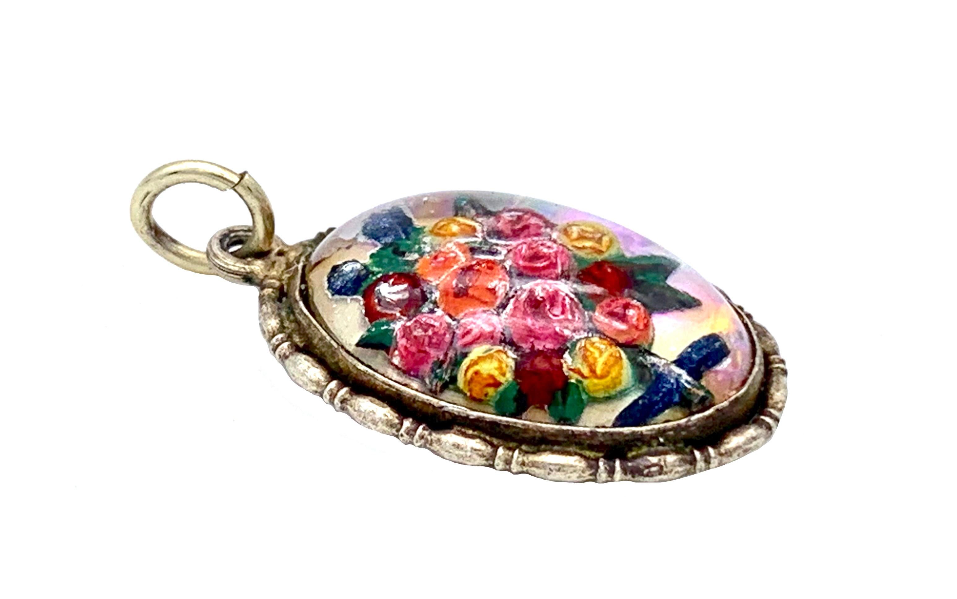 This charming little oval silver pendant is set with a cabochon shaped reverse painted crystal depicting a flower bouquet composed of red, pink, orange and yellow roses and green leaves.
The ornamental silver mount is typical for 1910/12. 
The