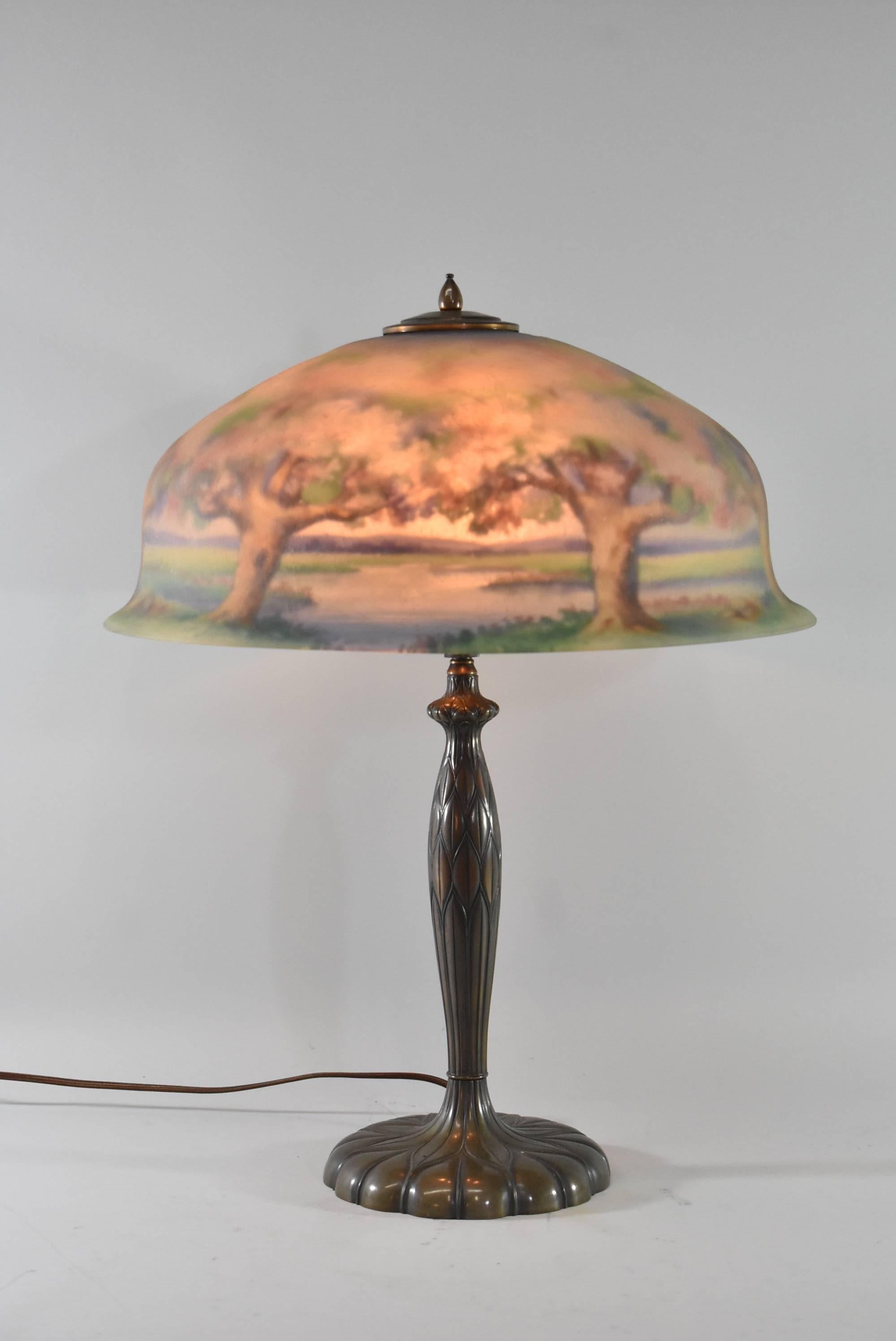 A beautiful reverse painted Pairpoint lamp signed by artist, W. Macy. This stunning lamp features three sockets and the shade depicts a forest scene in shades of blue, pink and green. The dimensions are 23