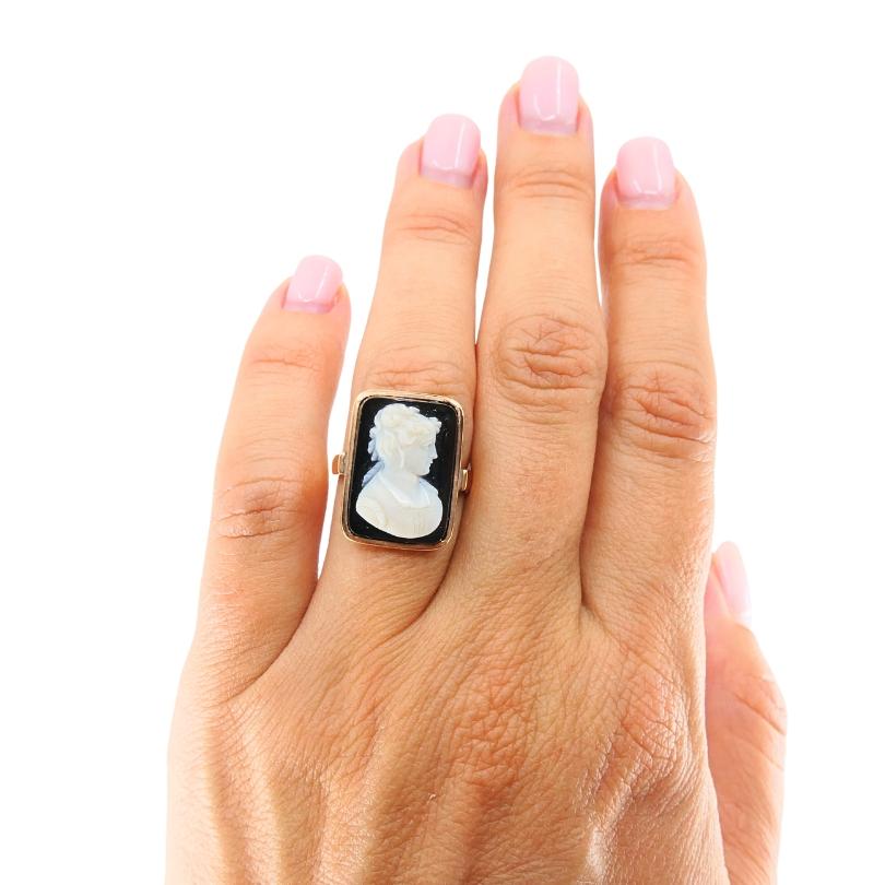 Questo anello antico è realizzato in oro 14k e presenta un cammeo rettangolare al centro. Il cammeo è scolpito in rilievo da una pietra stratificata, con un ritratto bianco di una donna su uno sfondo scuro. La figura è dettagliata con