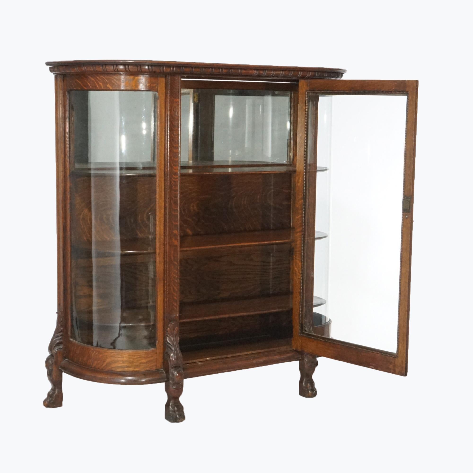 An antique China cabinet in the manner of RJ Horner offers quarter sawn oak construction in demilune form with single glass door opening to mirrored and shelved interior having flanking curved glass sides, raised on carved paw feet having acanthus