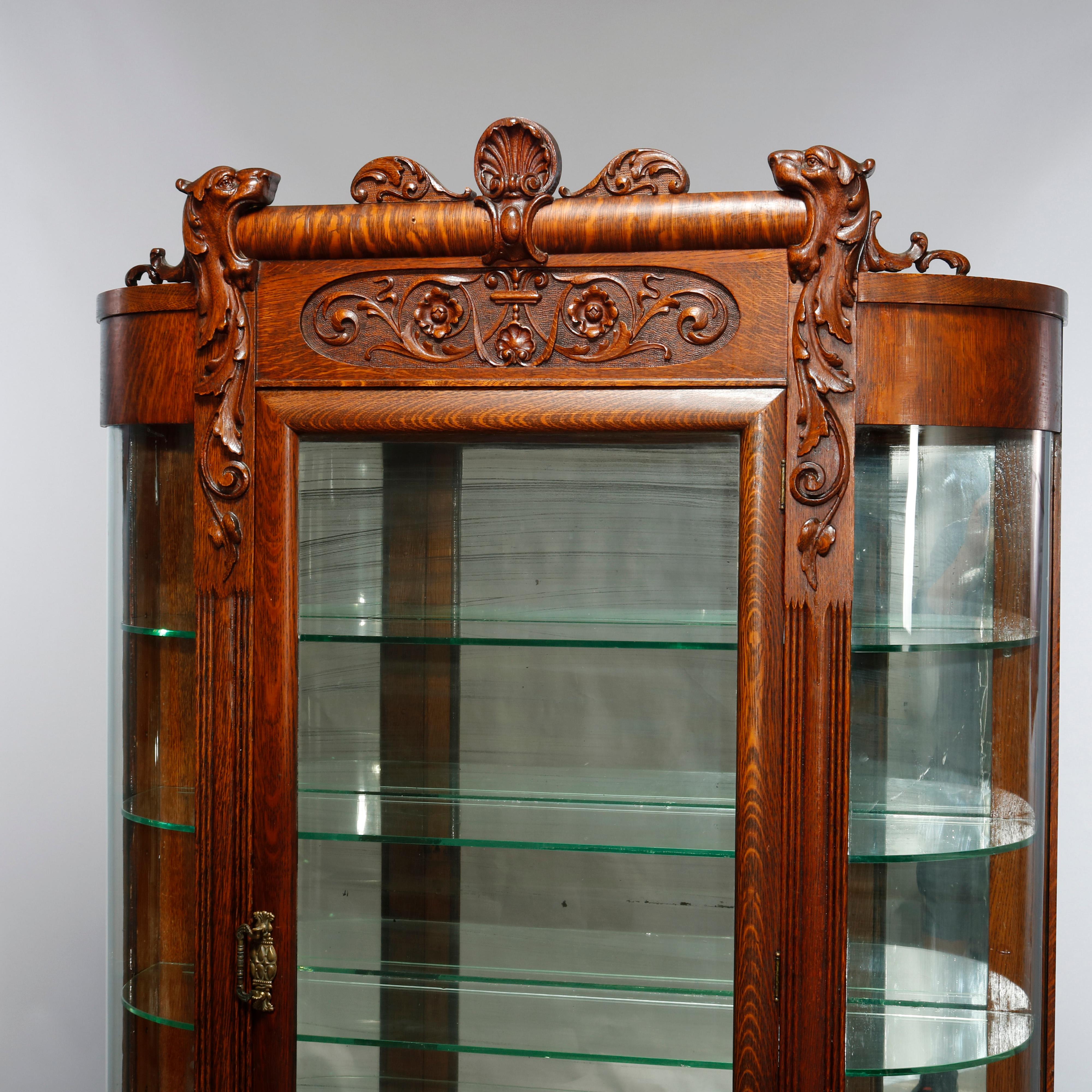 An antique china cabinet in the manner of RJ Horner offers oak construction with foliate cared crest having central carved shell and deeply striated quartersawn frieze surmounting case having curved glass sides and single door opening to shelved and