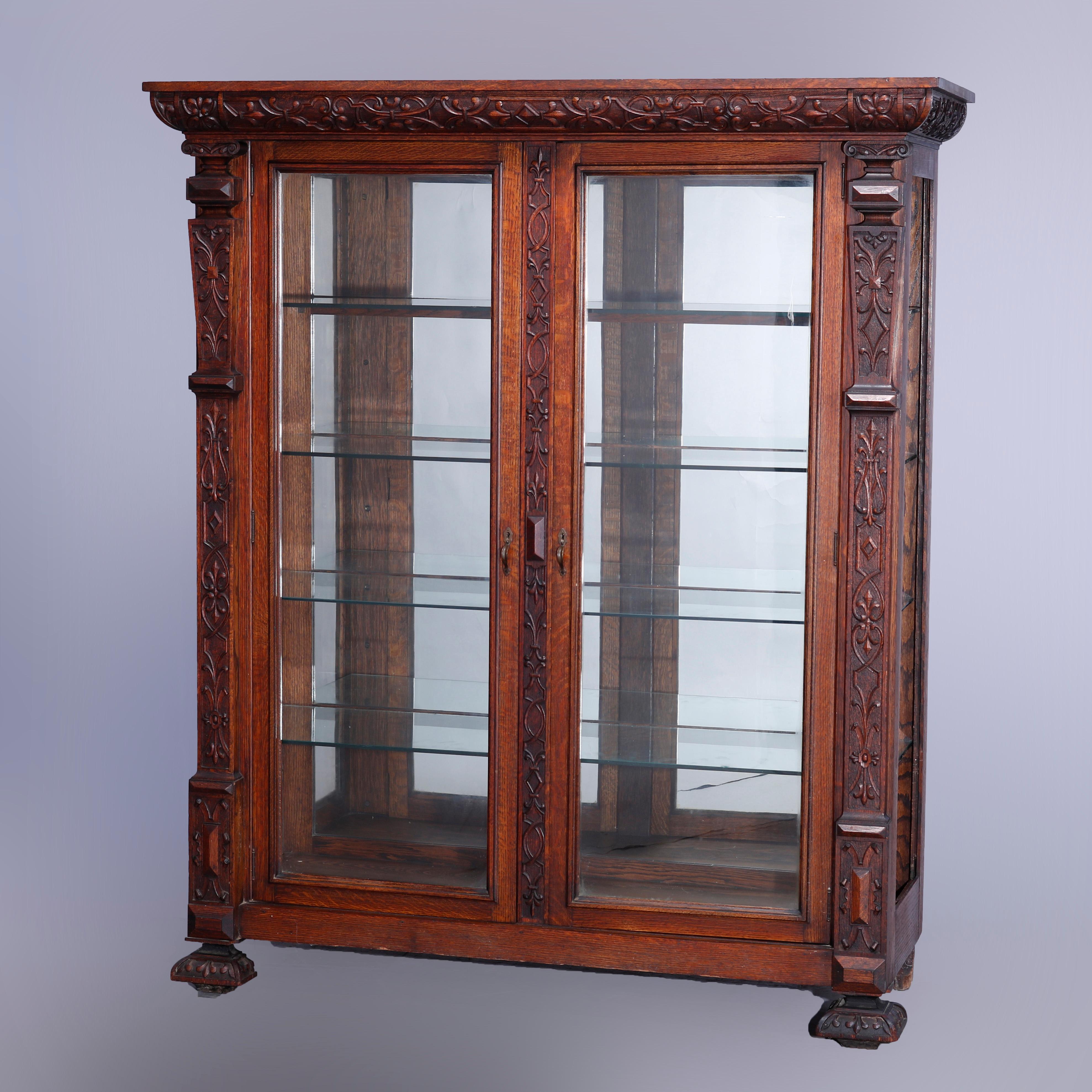 An antique enclosed china cabinet in the manner of RJ Horner offers quarter sawn oak construction with scroll and foliate carving throughout, double glass doors opening to glass shelved and mirrored interior, raised on foliate carved stylized bun