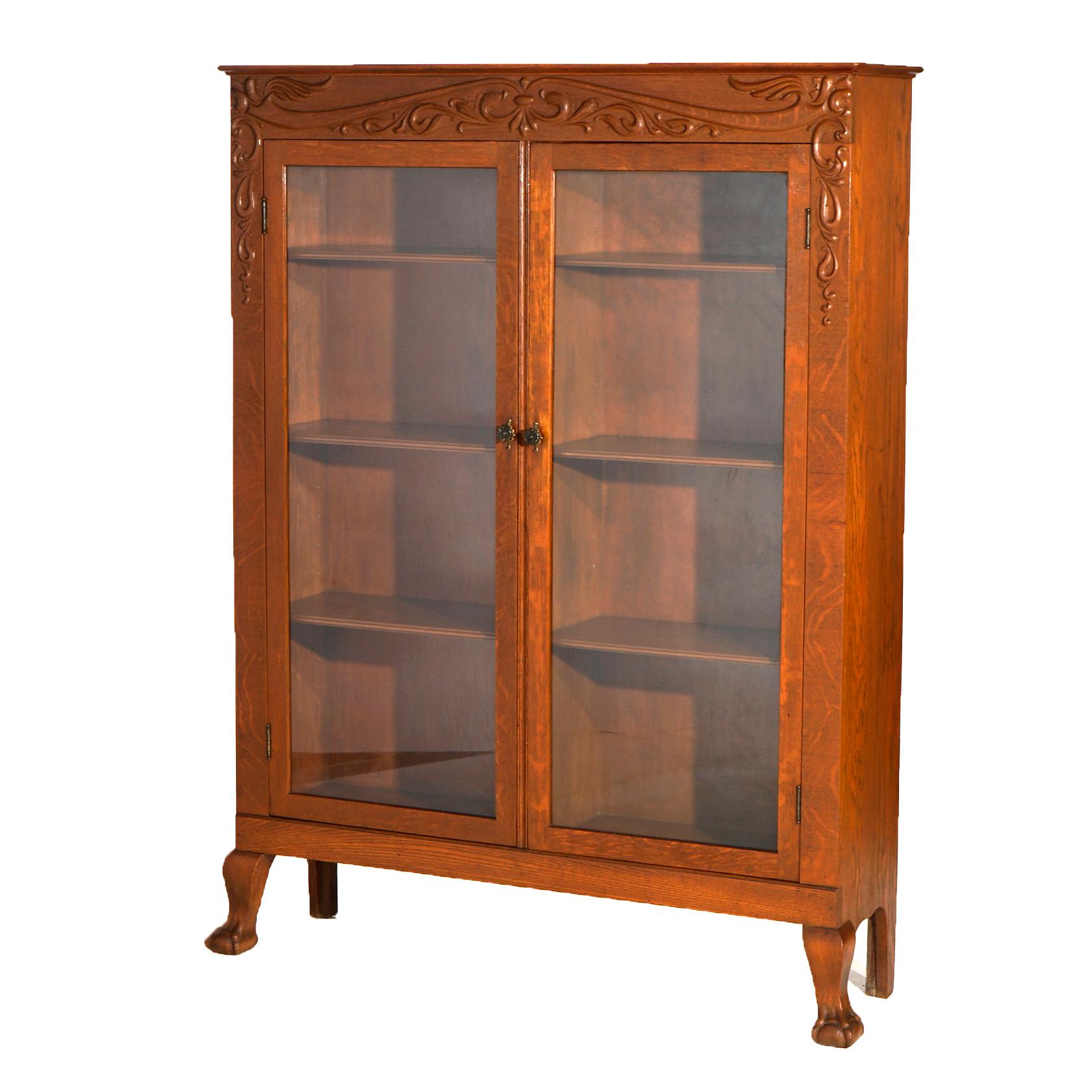 An antique bookcase in the manner of RJ Horner offers quarter sawn oak construction with frieze having scroll and foliate decoration over case having double glass doors opening to dived shelved interior, raised on cabriole legs terminating in claw