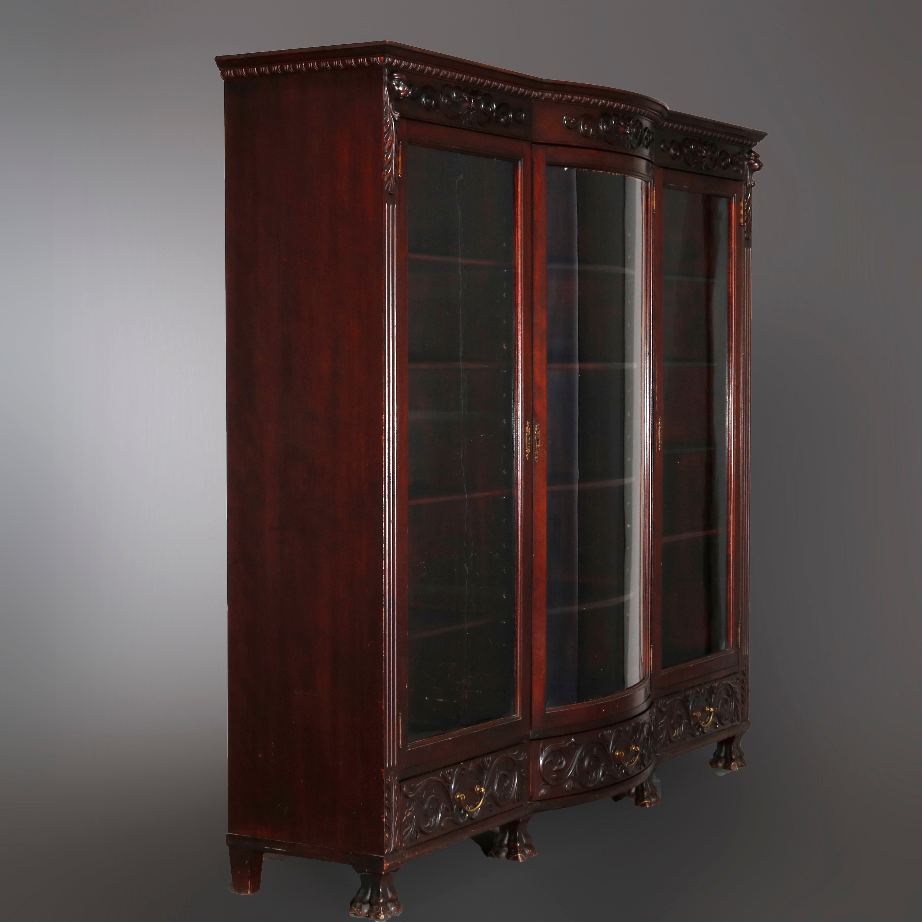 An antique figural bookcase in the manner of R.J. Horner offers mahogany construction with central bow front glass door flanked by single door bookcases and foliate carved crest with lion heads, over storage drawers and raised on carved paw feet,