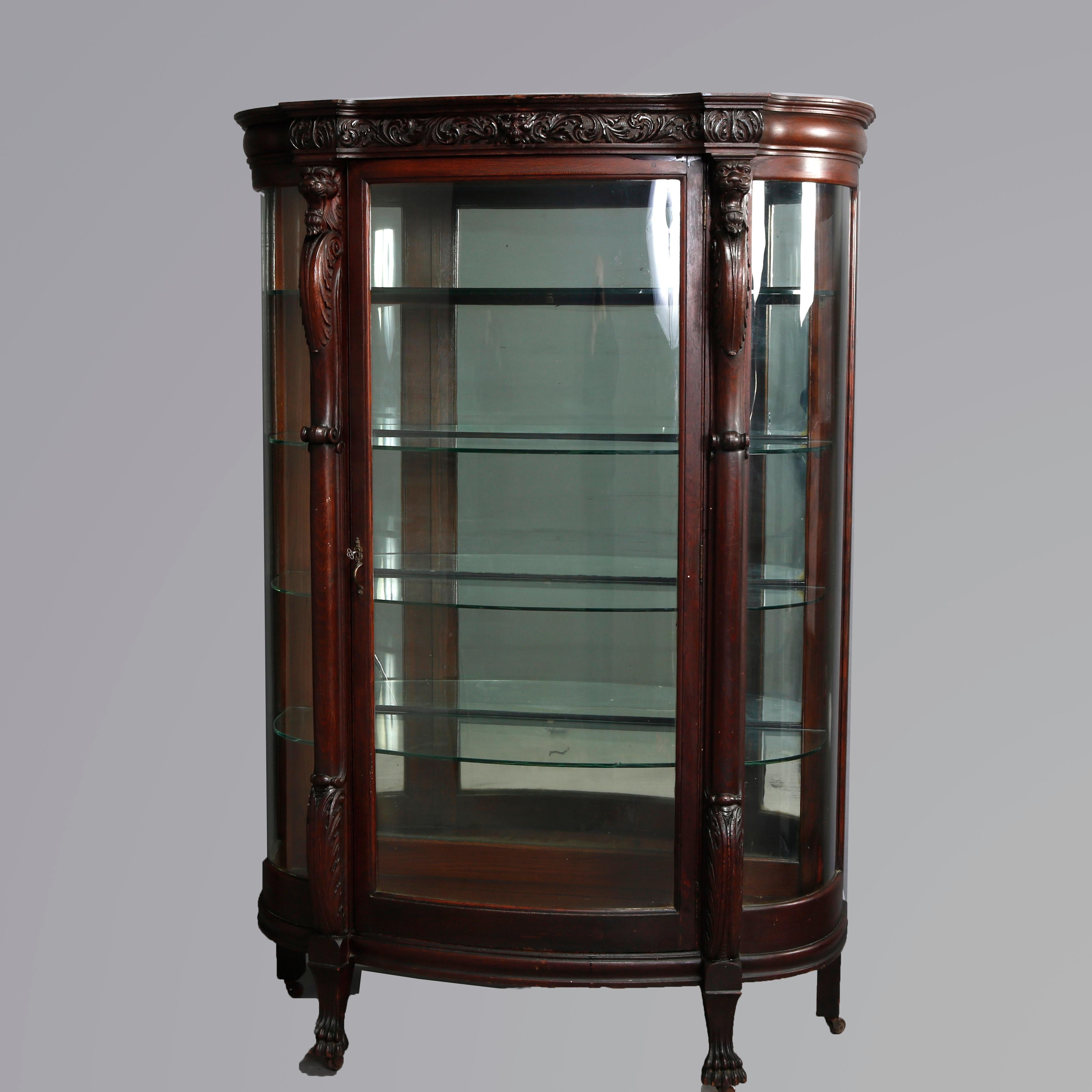 An antique R.J. Horner School oak china cabinet offers oak construction with carved wind god and foliate scrolled frieze surmounting case with lion heads flanking bent glass door, glass shelved and mirrored interior and carved acanthus foliate