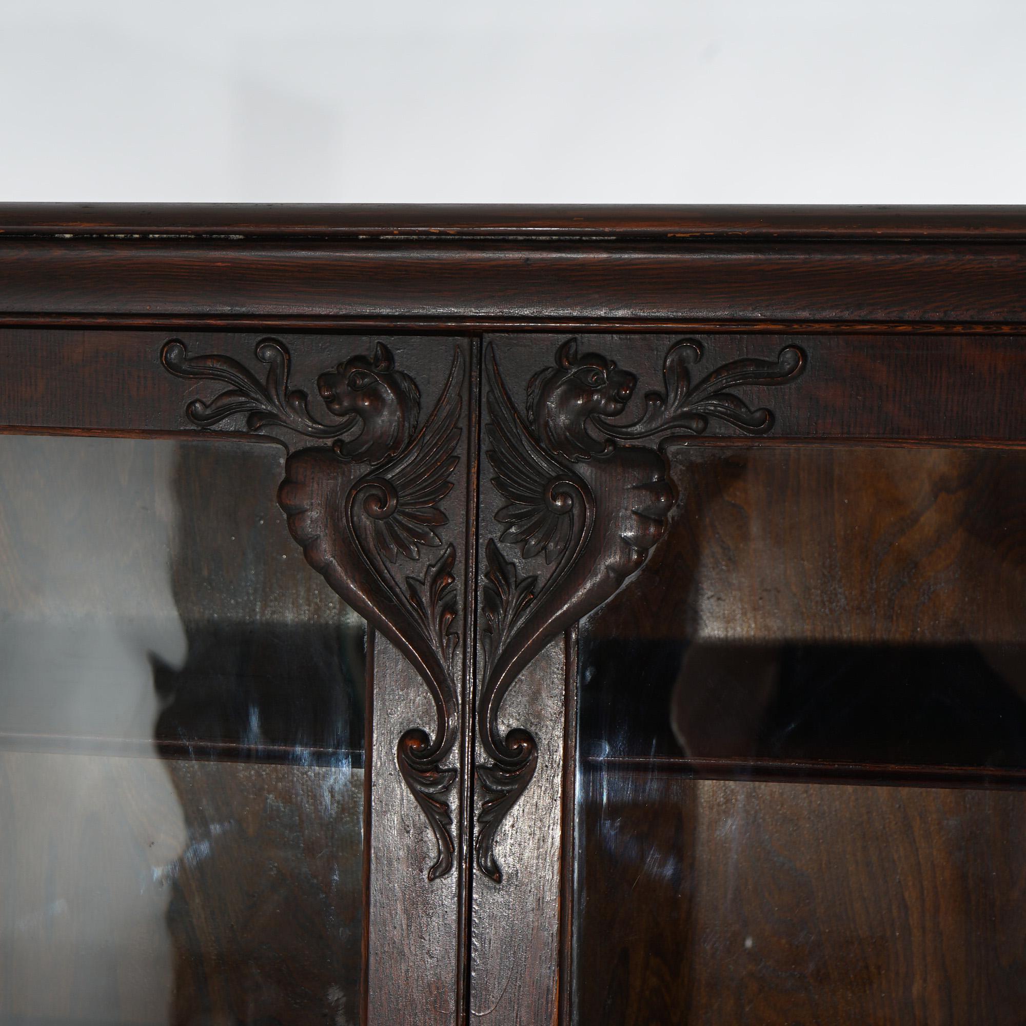 An antique bookcase in the manner of RJ Horner offers oak construction with carved griffins and foliate elements, double glass doors opening to divided and shelved interiors, raised on bracket feet, c1910

Measure - 60