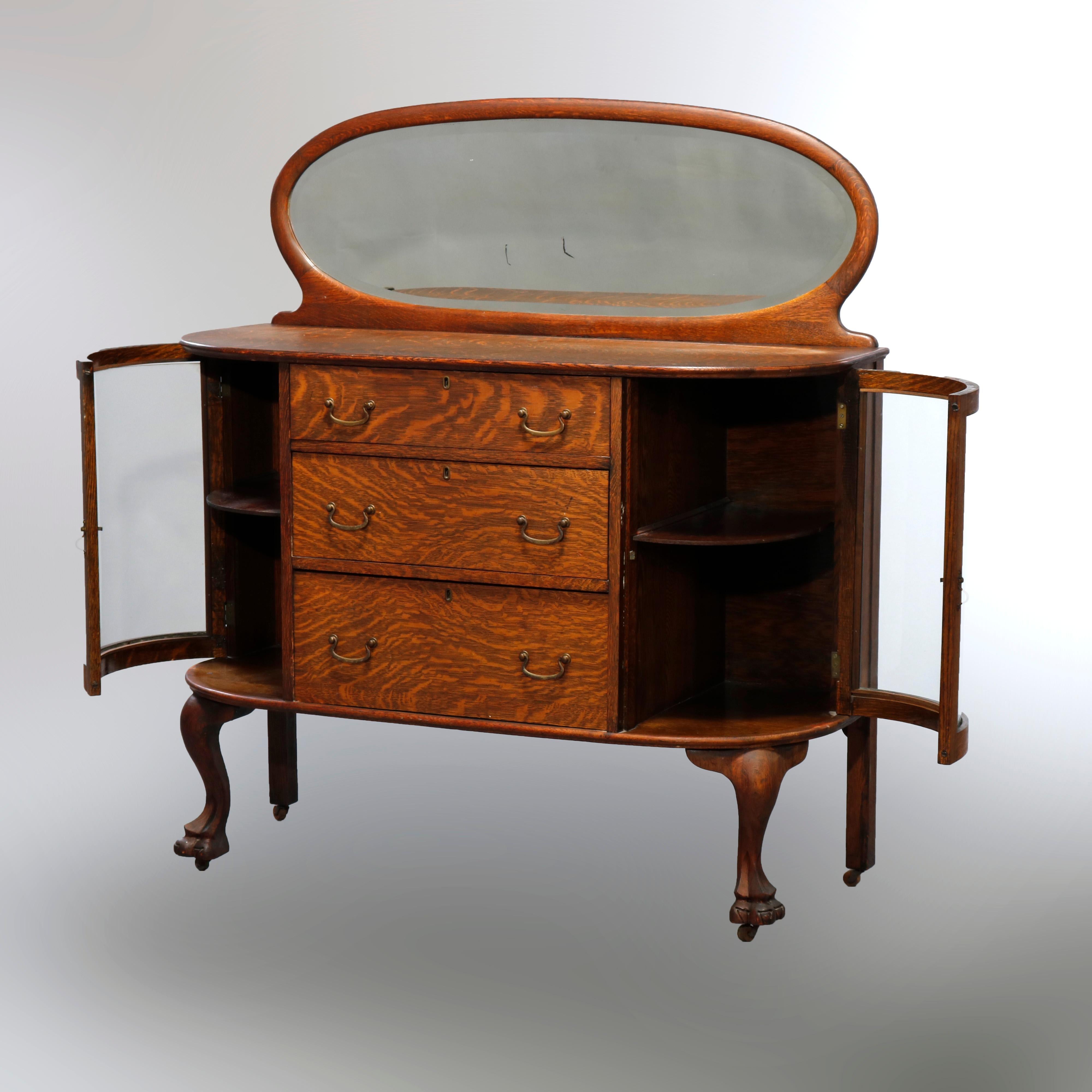 An antique china buffet in the manner of RJ Horner offers quarter sawn oak construction with an oval beveled mirror surmounting demilune case with central drawer tower having flanking display shelves and raised on cabriole legs terminating in carved