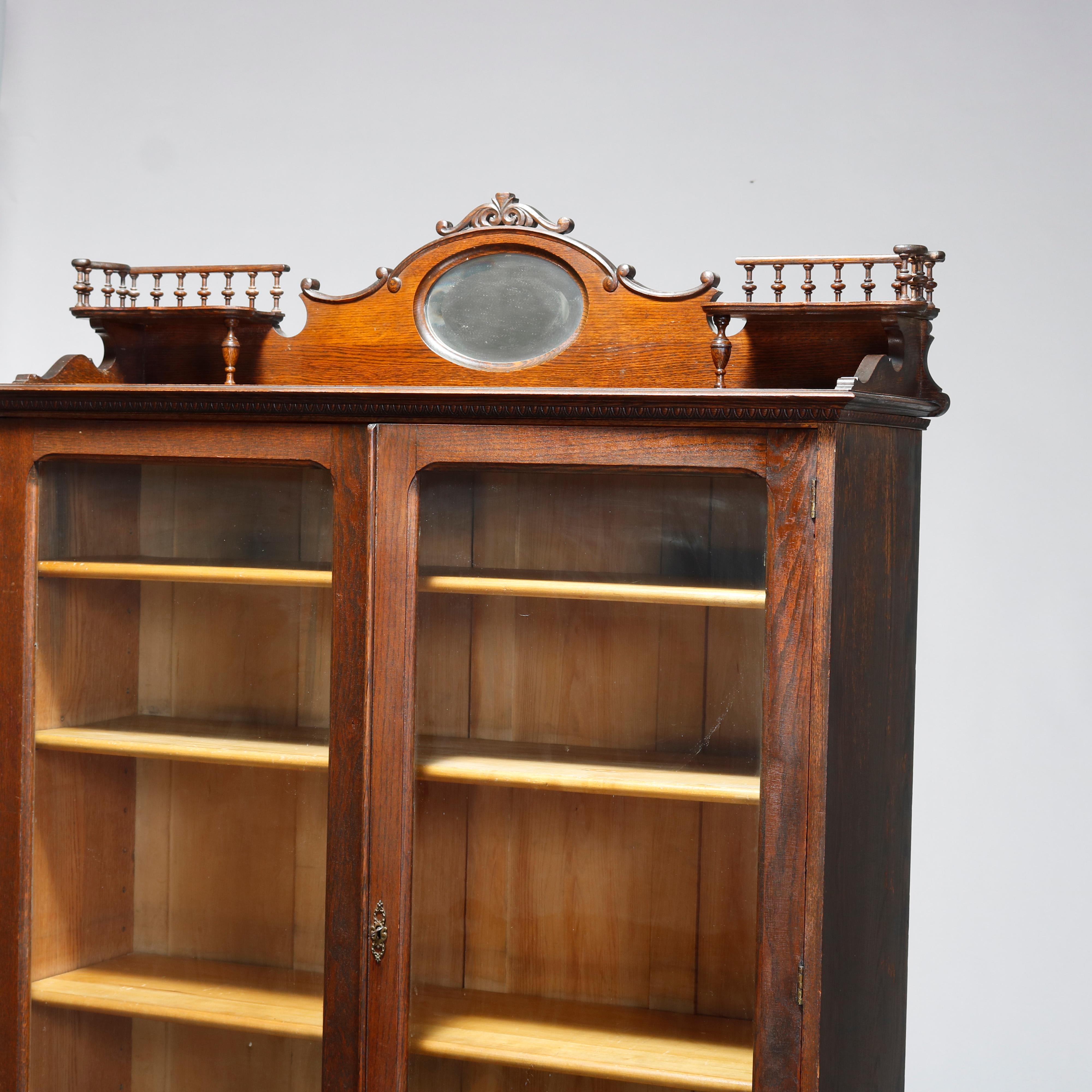 An antique bookcase by Sheboygan and in the manner of RJ Horner offers oak construction with shaped and mirrored backsplash having foliate carved crest with flanking spindle corner galleries surmounting double glass door bookcase with lower drawers,