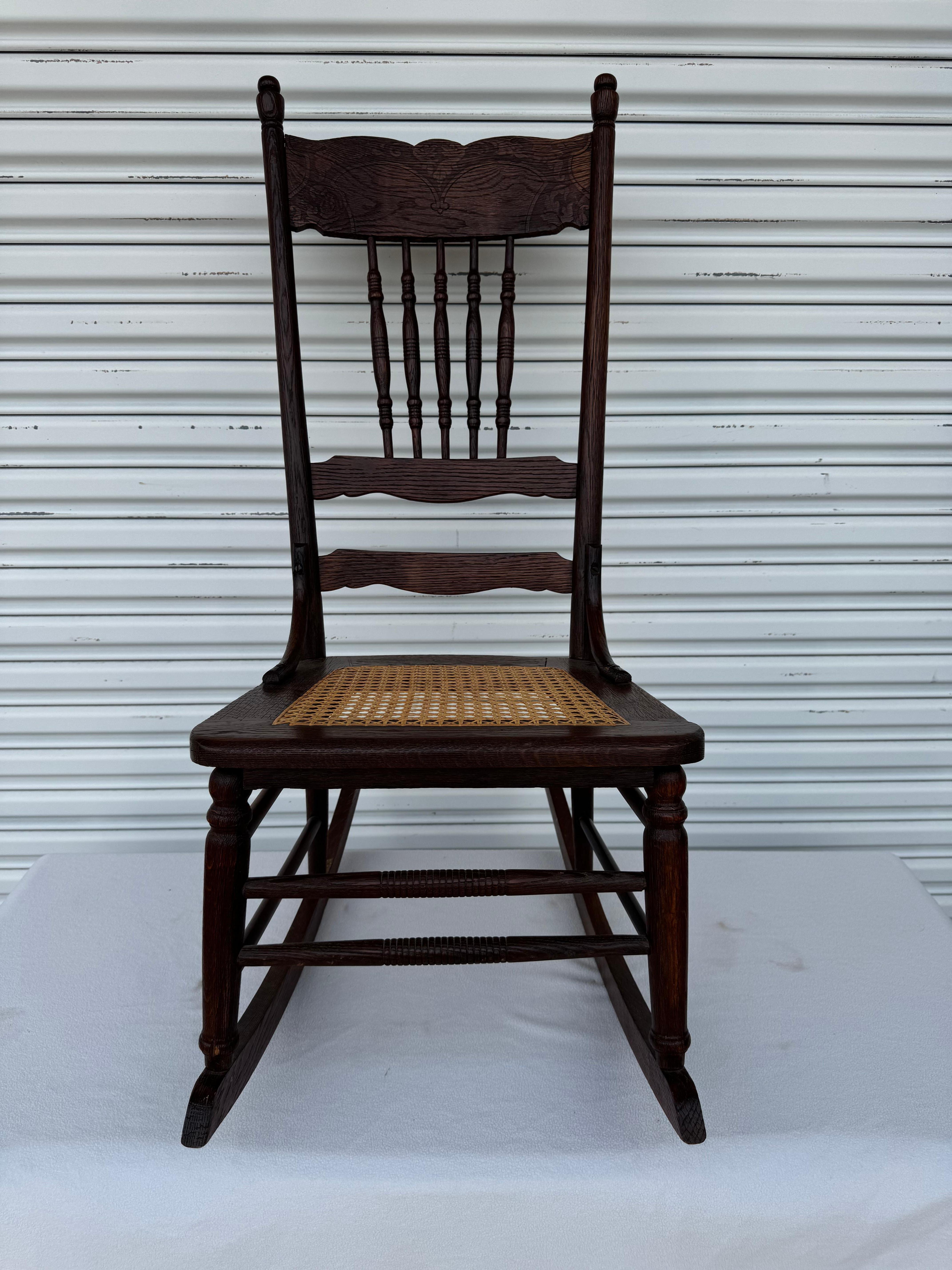 Antica sedia a dondolo in rovere massiccio con seduta in canna in buone condizioni. Questa sedia è caratterizzata da una cresta smerlata e frondosa che sovrasta i fusi torniti. Si tratta di un bellissimo pezzo d'antiquariato che fa bella mostra di