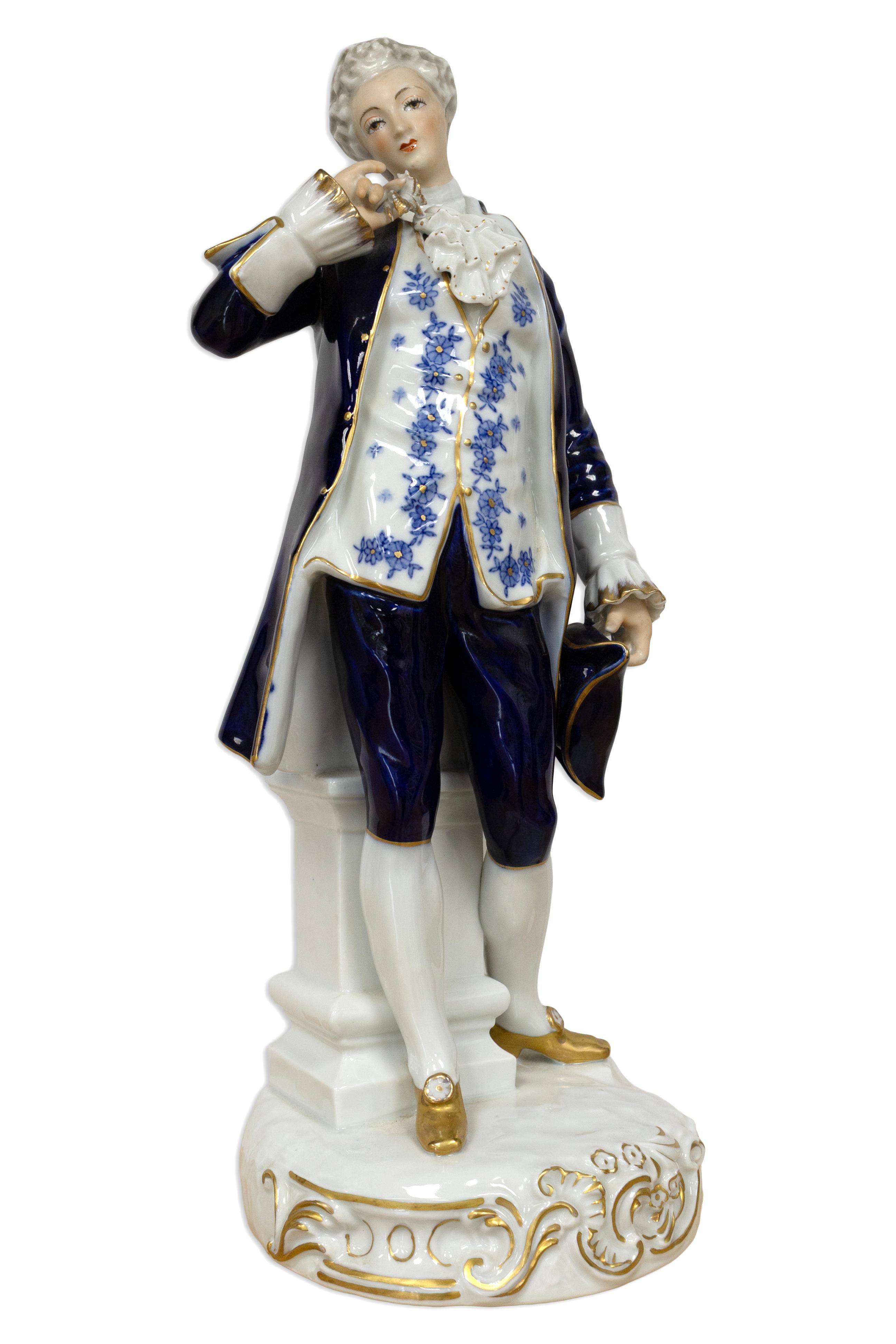 An elegant pair of German Rococo style painted porcelain figurines finely modeled as aristocratic man and woman in 18th-century court attire. The gentleman is depicted in a cobalt blue and white suit richly accented with floral motifs and gilt trim,