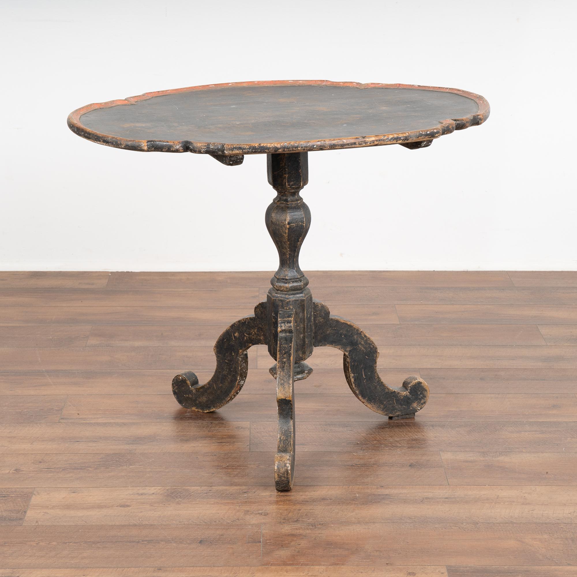 The aged patina of the original black paint accentuates the curves and grace of this Rococo tilt top table side table.
The black painted finish with red trim around the edge has a warm aged patina from generations of use, distressed to reveal the