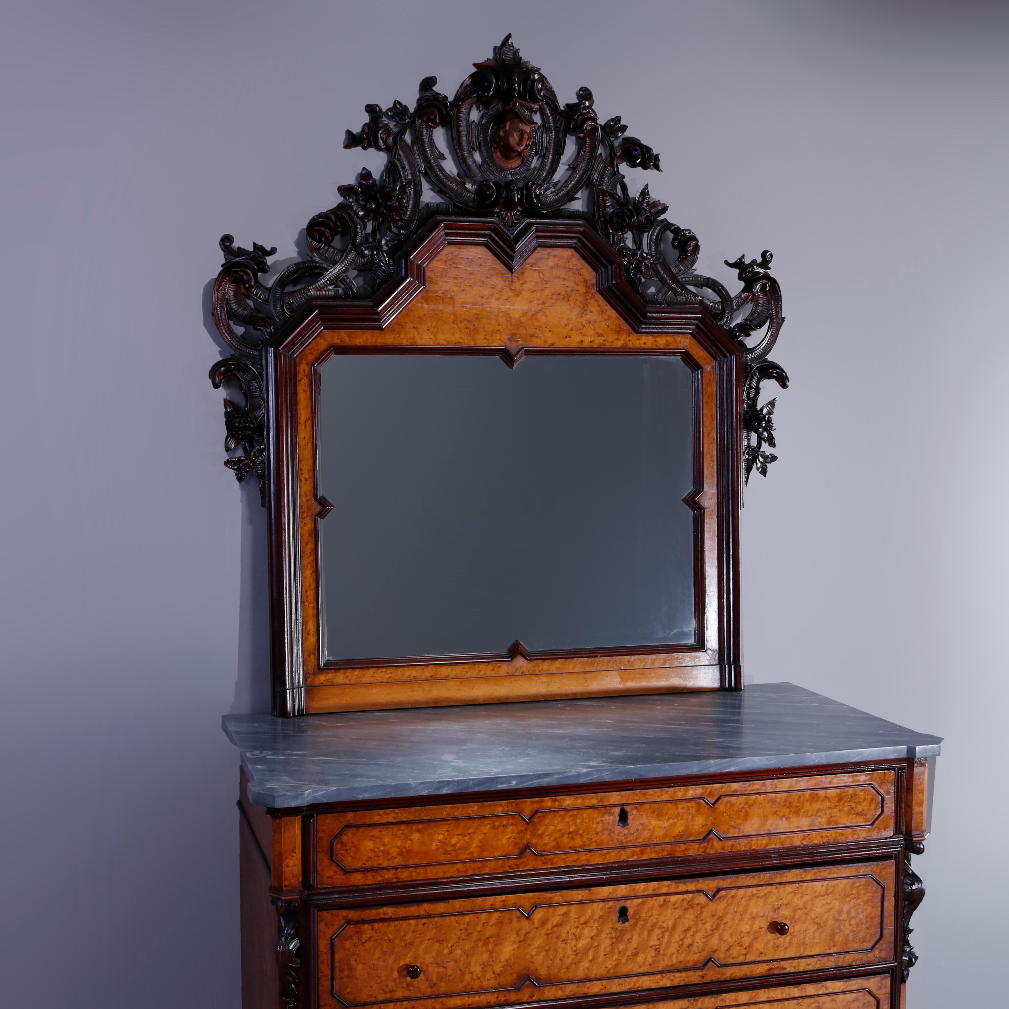 An antique Rococo Revival figural and mirrored chest of drawers offers birdseye maple construction with heavily carved and pierced foliate walnut crest with central mask over mirror surmounting lower marble top case with paneled long drawers with