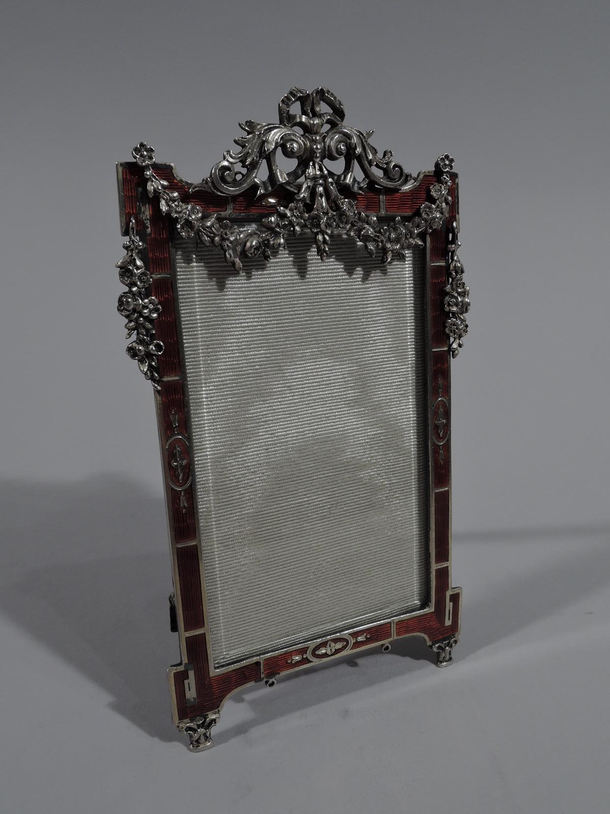 Rococo Revival silver and enamel picture frame, ca 1910. Rectangular window. Surround flat block volute corners. Dramatic red guilloche enamel and inlaid stylized leaves. Applied ribbon and leaf crown with overhanging flower garland. Two capital