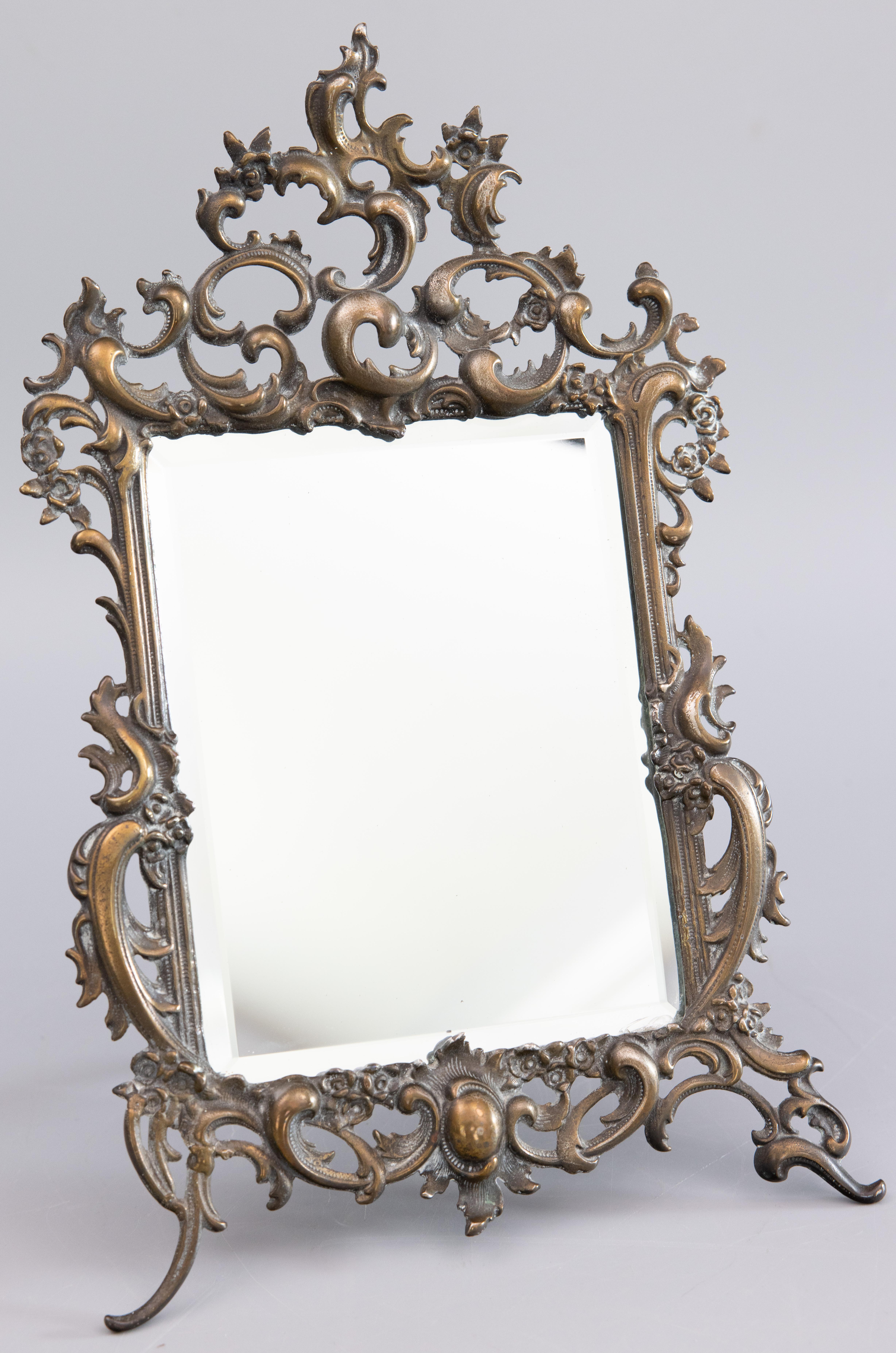 A gorgeous antique French Rococo style brass tabletop dresser or vanity easel back mirror with an ornate scroll border, circa 1900. This fine mirror is well made and heavy, weighing 4 lbs, with a lovely patina and it retains the original beveled