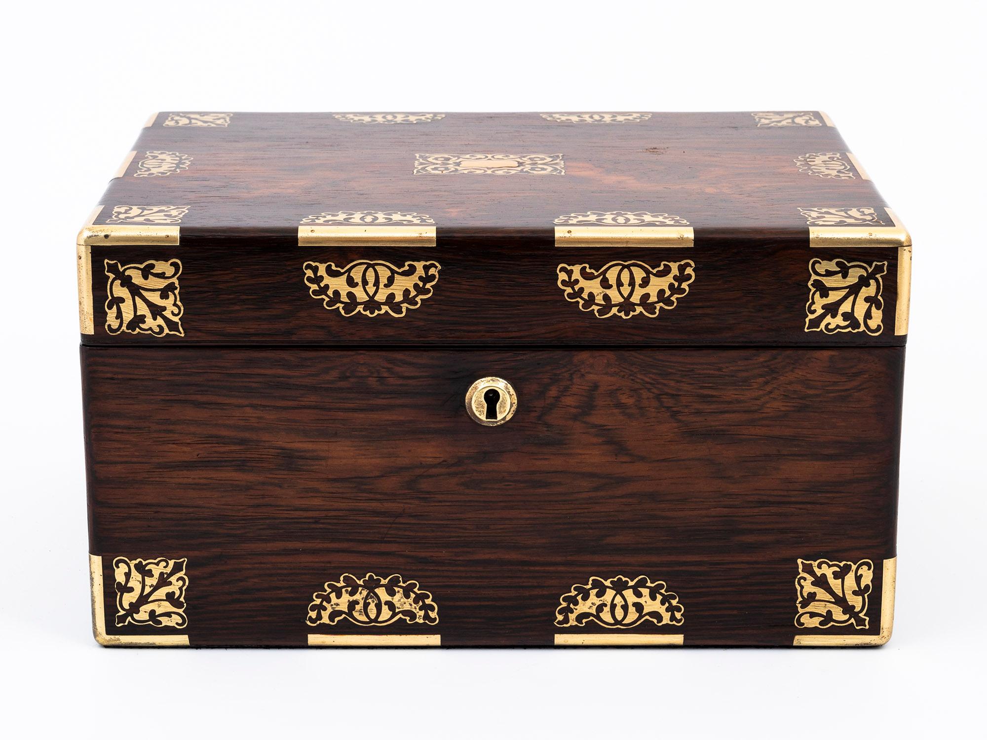 The following is a truly remarkable Antique Rosewood Jewellery Box, boasting an unparalleled combination of style and practicality.

The top and front of the box are adorned with ornate Brass inlay, featuring segmented edging with a stunning